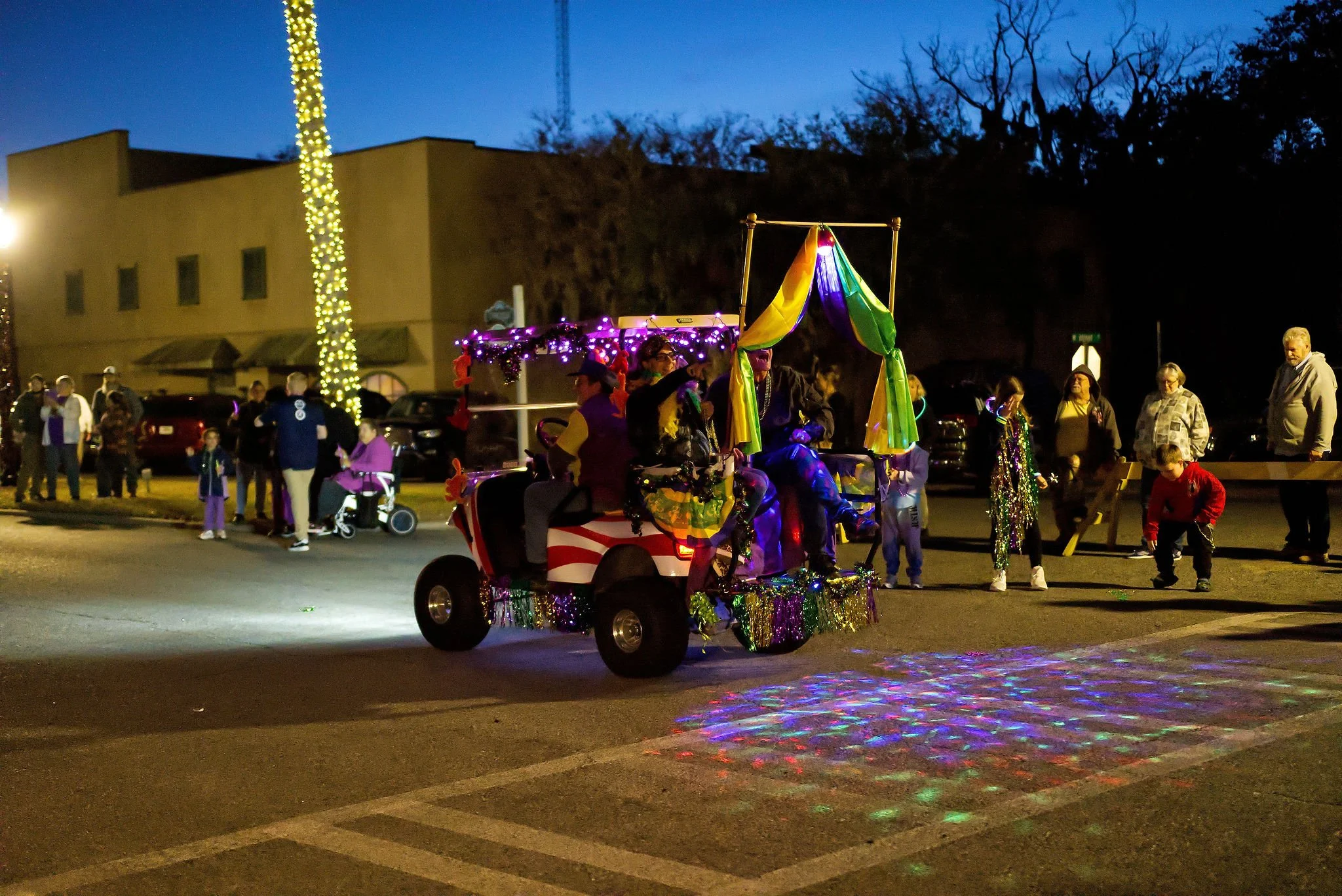 A decorated golf cart with shiny multicolored ribbons and lights, moving through a street during a nighttime parade, with spectators on the sidewalk watching.