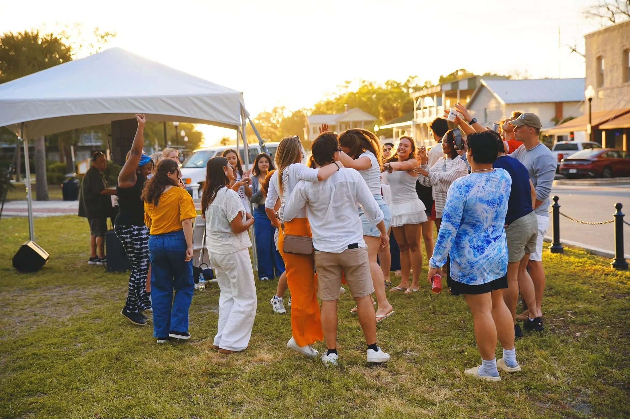 Group of people celebrating outdoors under a white canopy, captured during sunset, in a grassy area with houses and cars in the background.