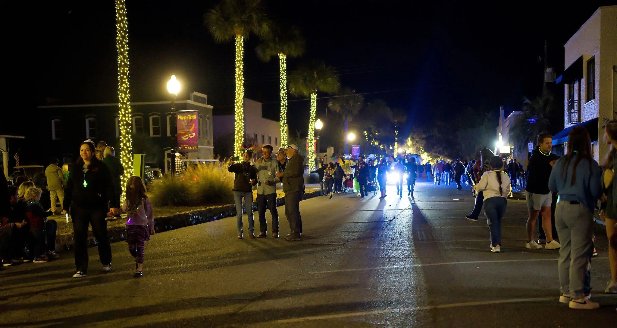 Nighttime street scene with people walking and talking, decorated with palm trees wrapped in yellow lights.
