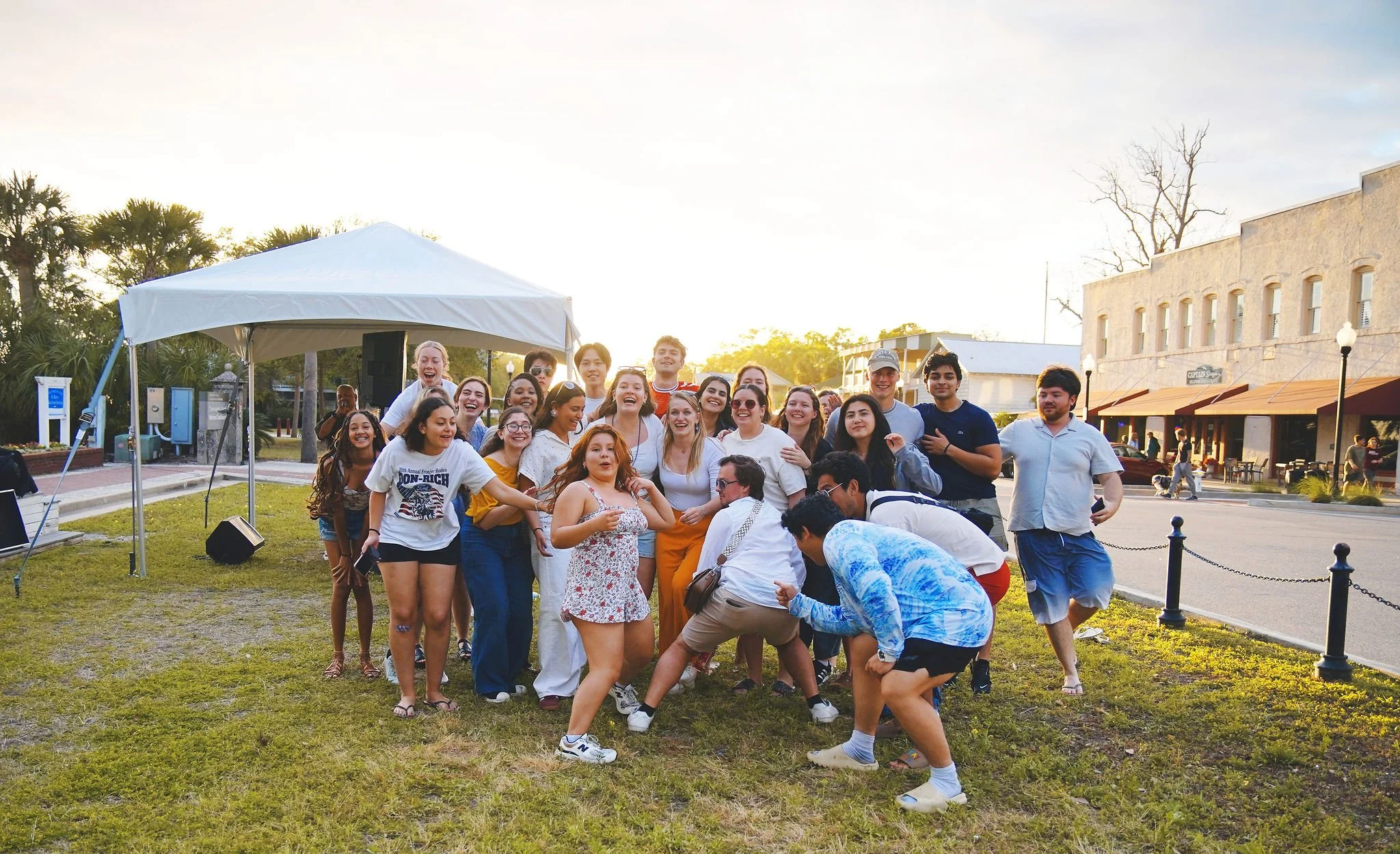 A group of young people gathered outdoors on a grassy area, smiling and posing for the photo with some making playful gestures. There is a white canopy tent and a row of buildings with shops and cafes in the background, during sunset.