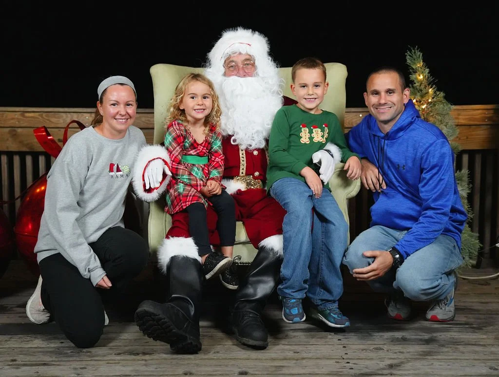 Family photo with Santa Claus, two children, and two adults, celebrating Christmas indoors, with a decorated Christmas tree in the background.