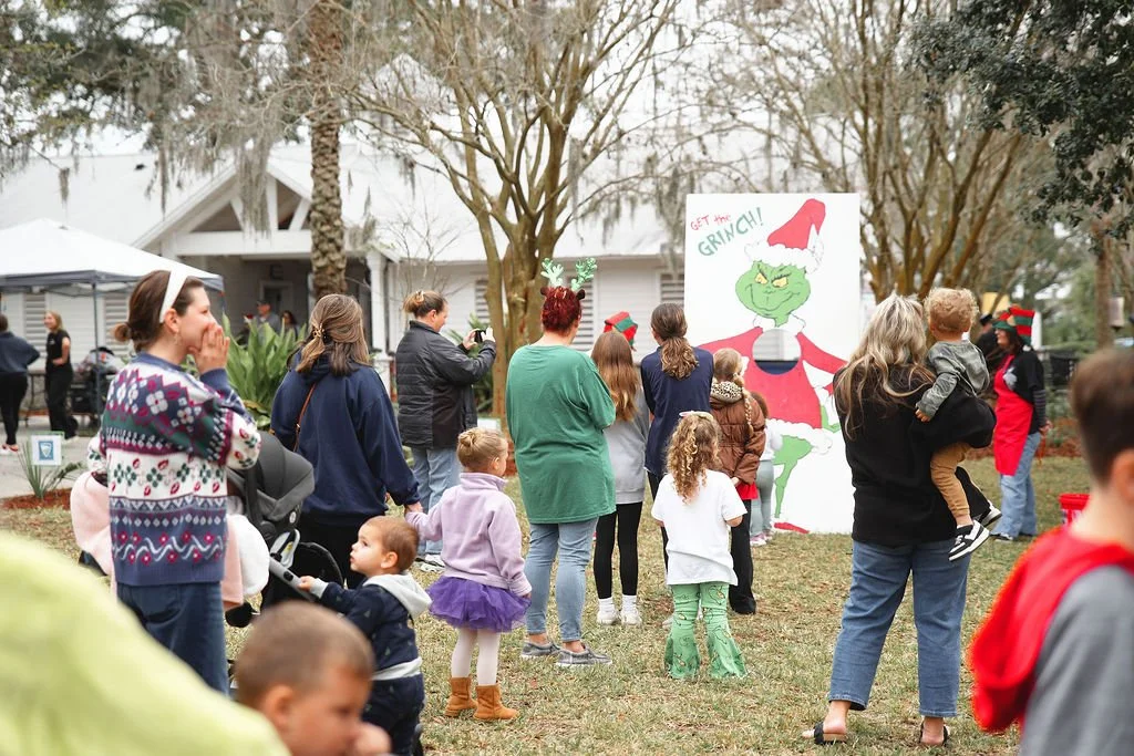 People gathered outdoors attending a Christmas event with a large Grinch-themed backdrop, trees, and festive decorations.