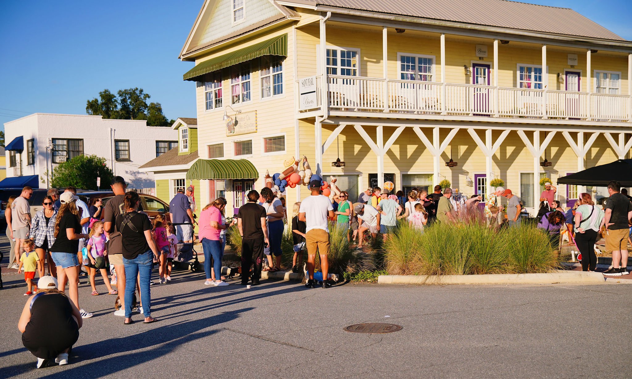 A crowd of people gathered outside a yellow, two-story building with white railings and a purple door, under a clear blue sky. The building has a green awning and balloon decorations. The scene suggests a community event or gathering.
