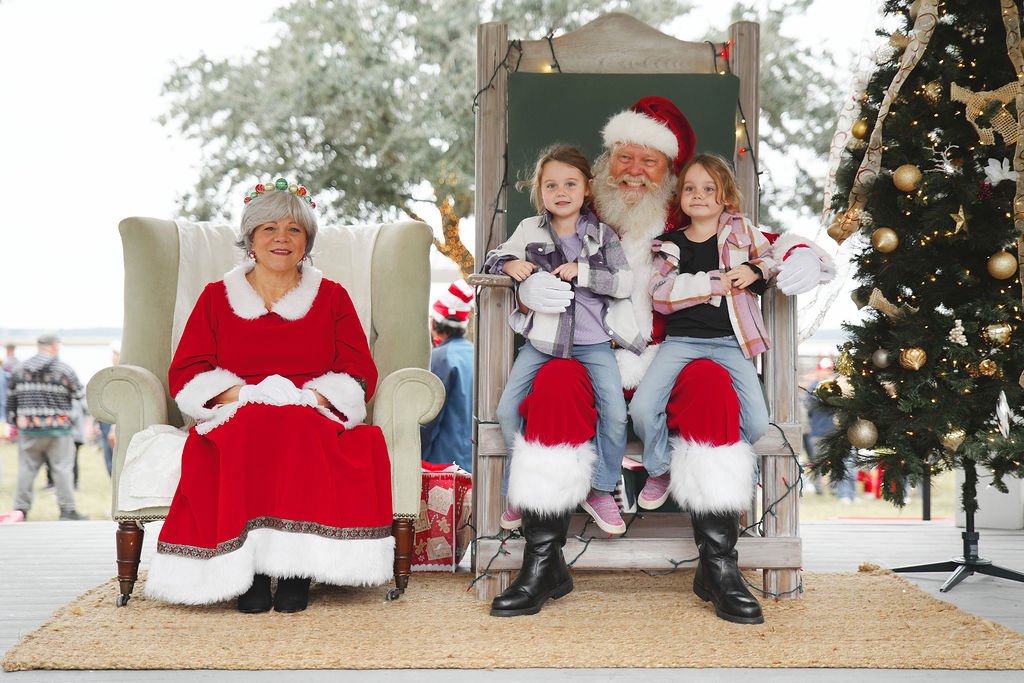 Santa Claus and two young girls sitting on a Christmas throne with a woman dressed as Mrs. Claus nearby, a decorated Christmas tree, and holiday background at an outdoor Christmas event.