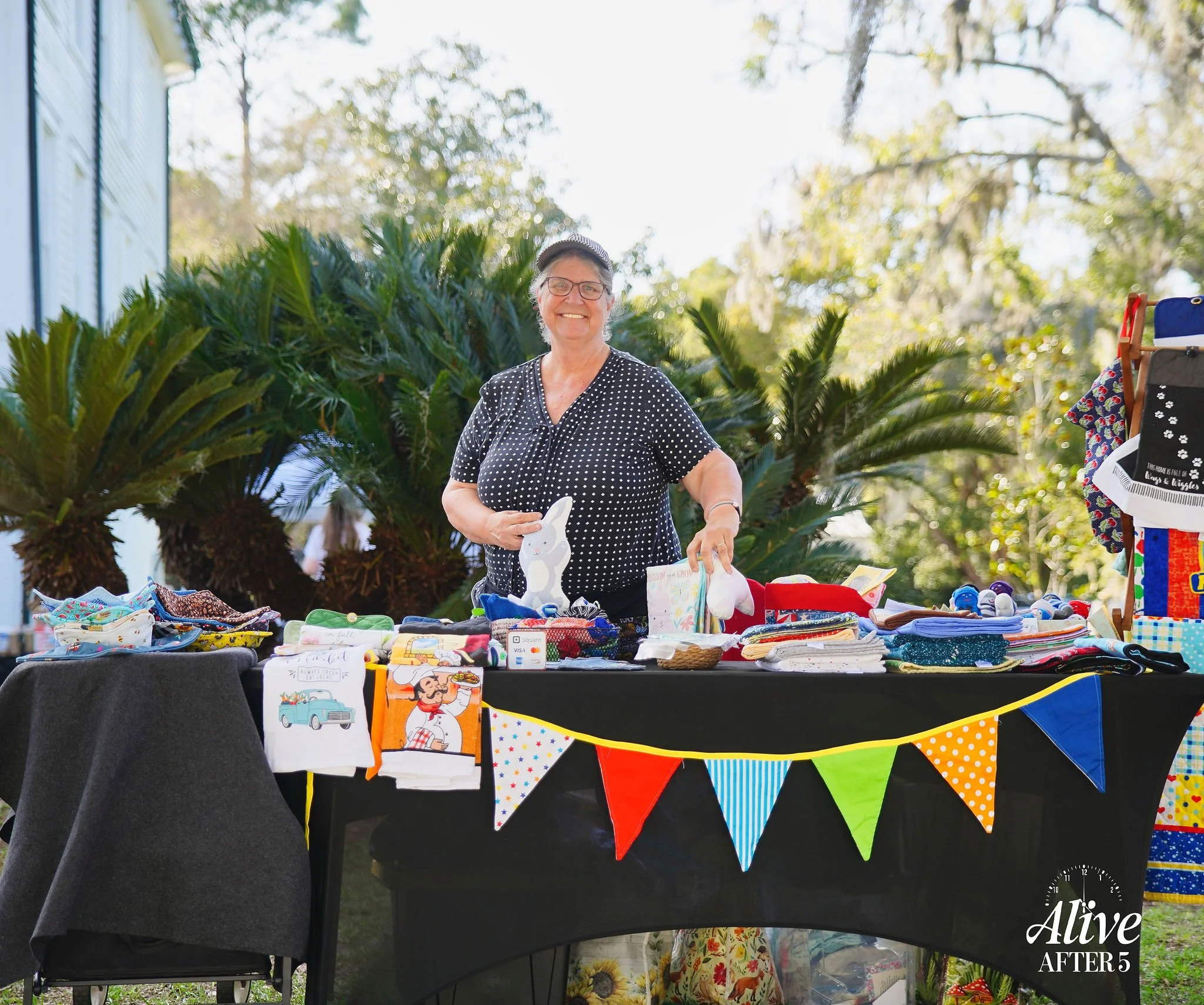 A smiling woman standing behind a table filled with various handcrafted items and fabric products, outdoors with trees in the background, at a community yard sale or craft fair.
