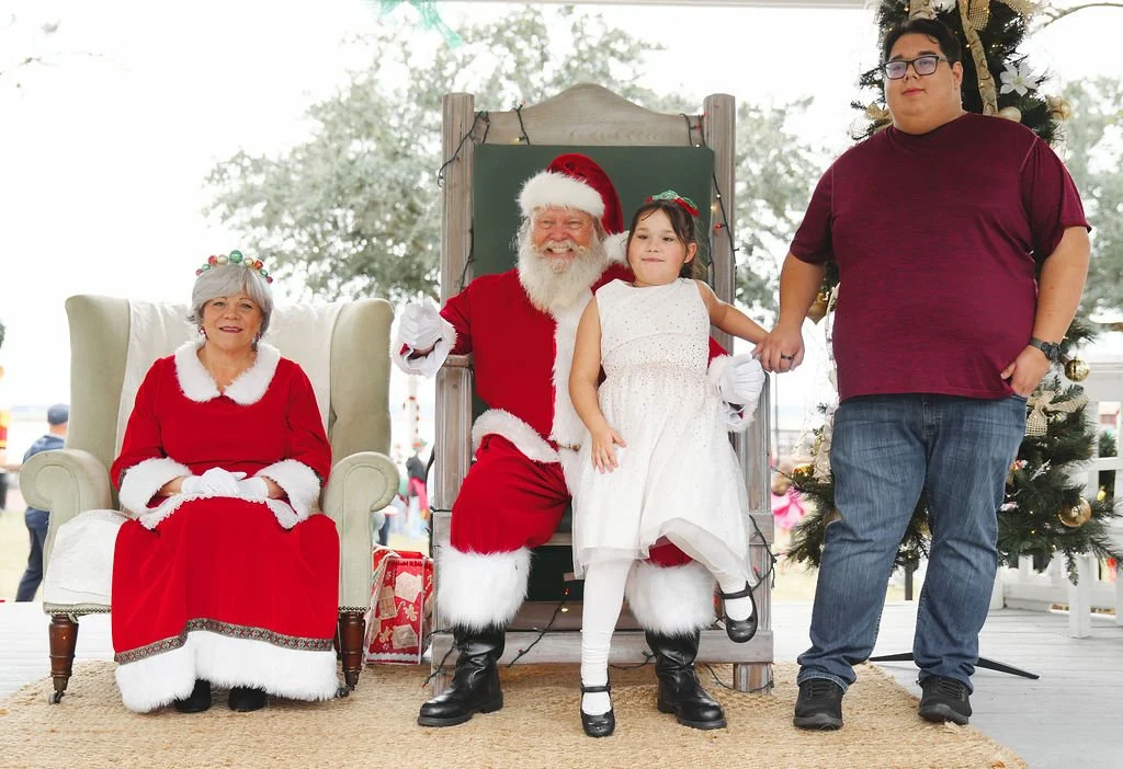 Family posing with Santa Claus at Christmas event, woman in Santa costume seated, man dressed as Santa with a girl in white dress on his lap, holding hands with boy in maroon shirt and jeans, decorated Christmas tree nearby.