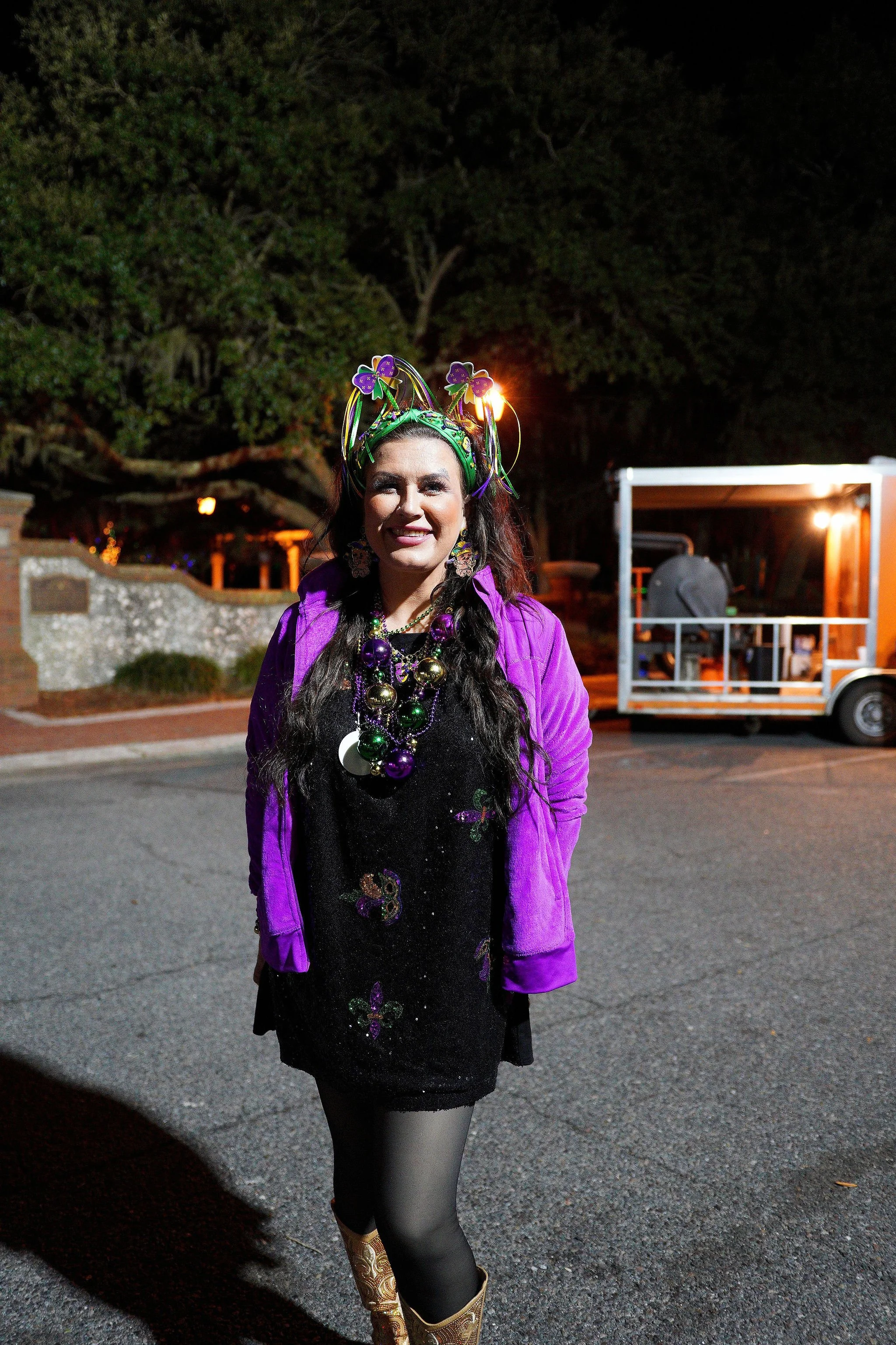 A woman dressed in purple and black, wearing Mardi Gras beads and a festive headpiece, standing outdoors at night with a lit trailer behind her.