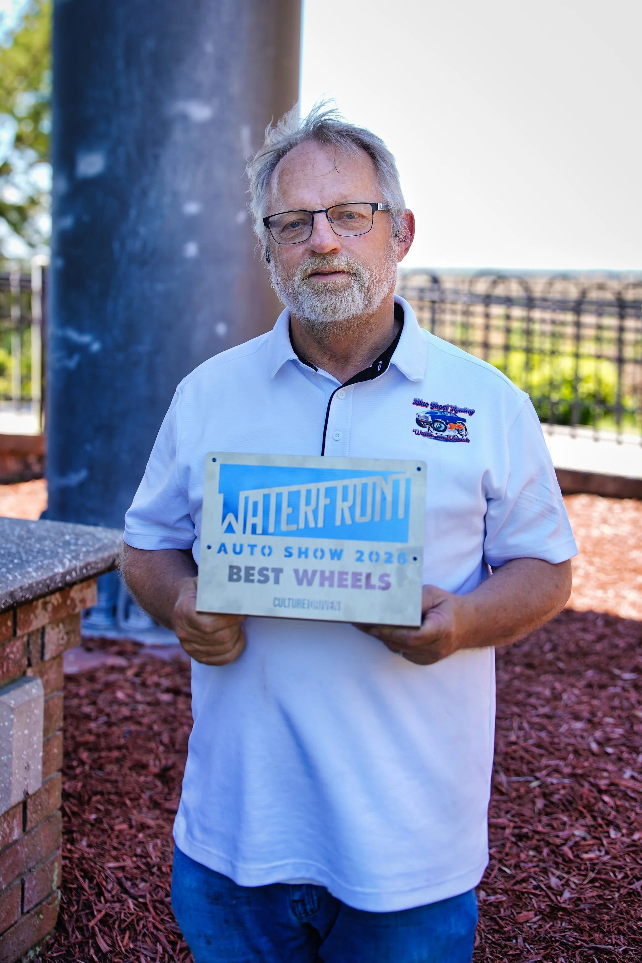 A man with glasses and gray hair holding an award plaque that reads 'Jalderf Run Auto Show 2026 Best Wheels.' He is standing outdoors in front of a large metal column and a fence, wearing a white collared shirt with a logo.