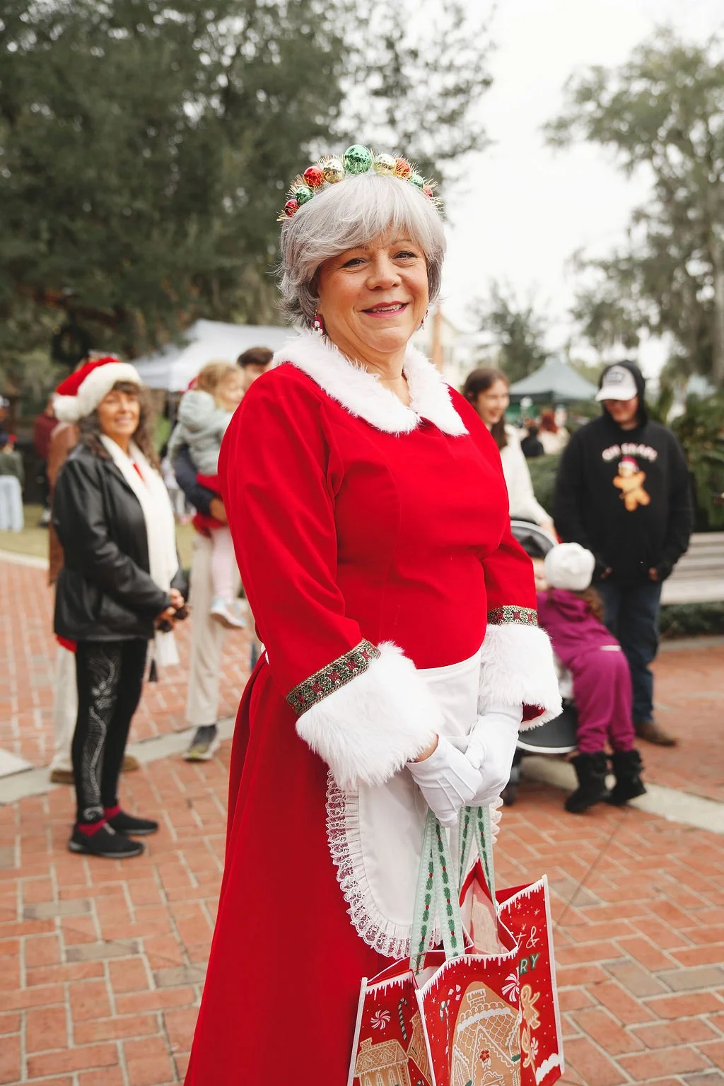 A woman dressed as Mrs. Claus in a red costume with white fur trim, wearing white gloves and carrying holiday shopping bags, standing outdoors at a festive event with other people in the background.