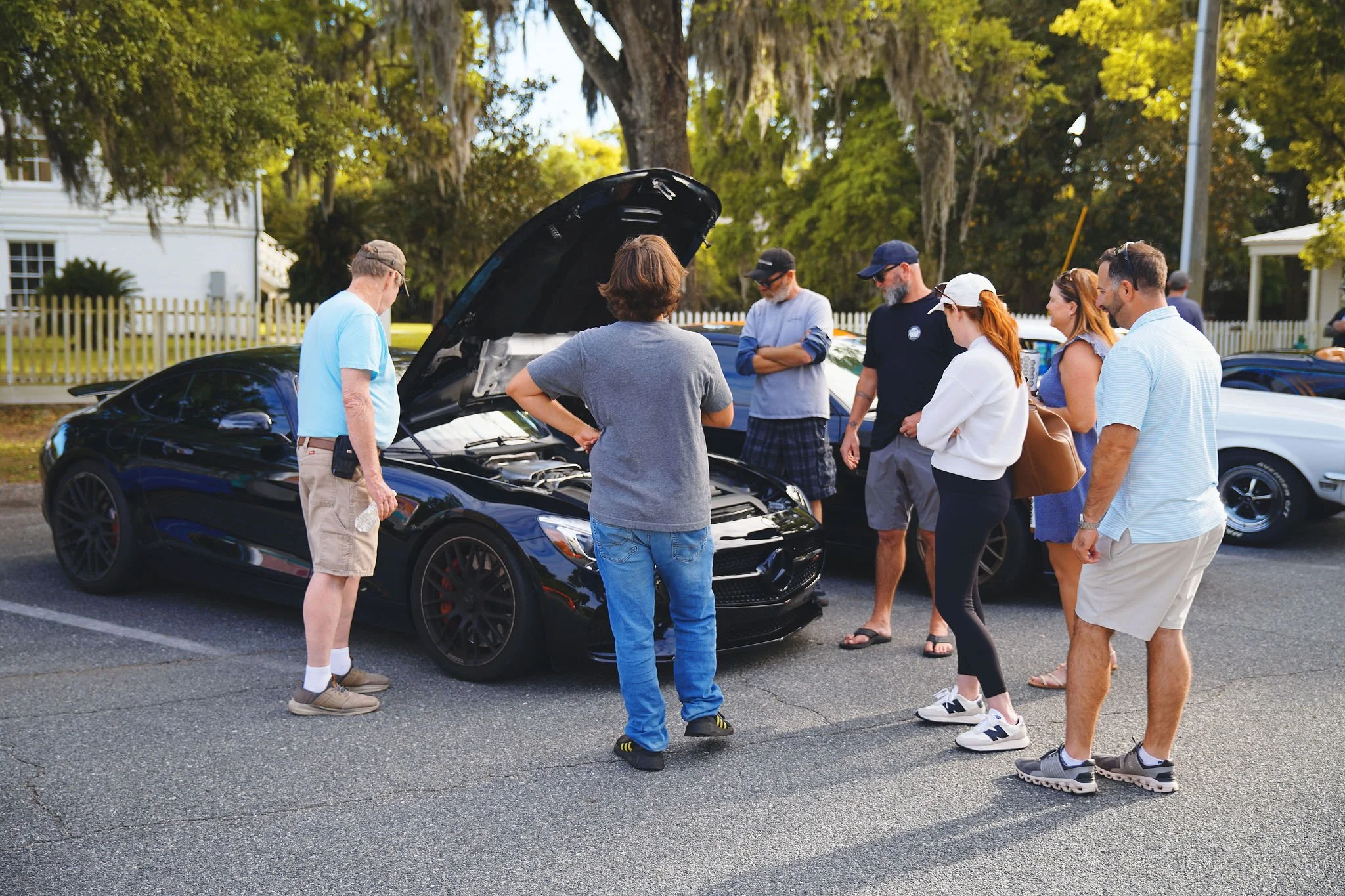 A group of people gathered around a black sports car with the hood open, looking at the engine, in a parking lot on a sunny day.