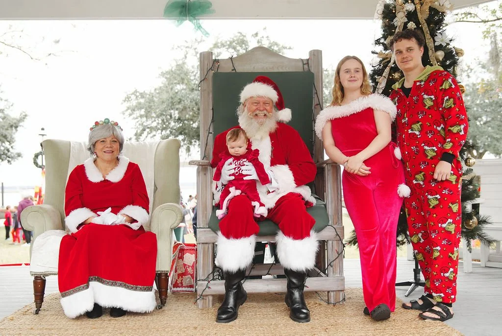 People dressed in Christmas pajamas and costumes, including Santa Claus with a baby, sitting on a Christmas-themed set with a decorated tree in the background.
