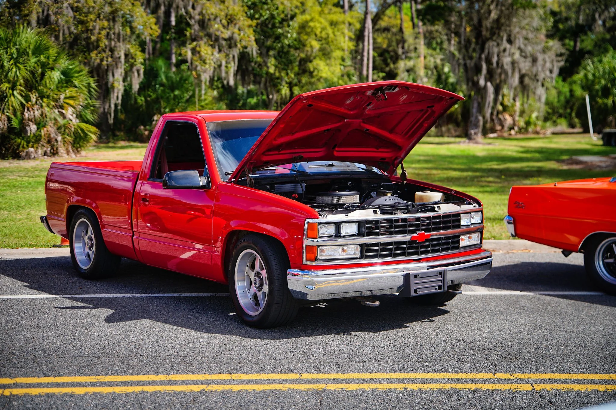 A red Chevrolet pickup truck with an open hood parked on a street, with green trees and grass in the background.