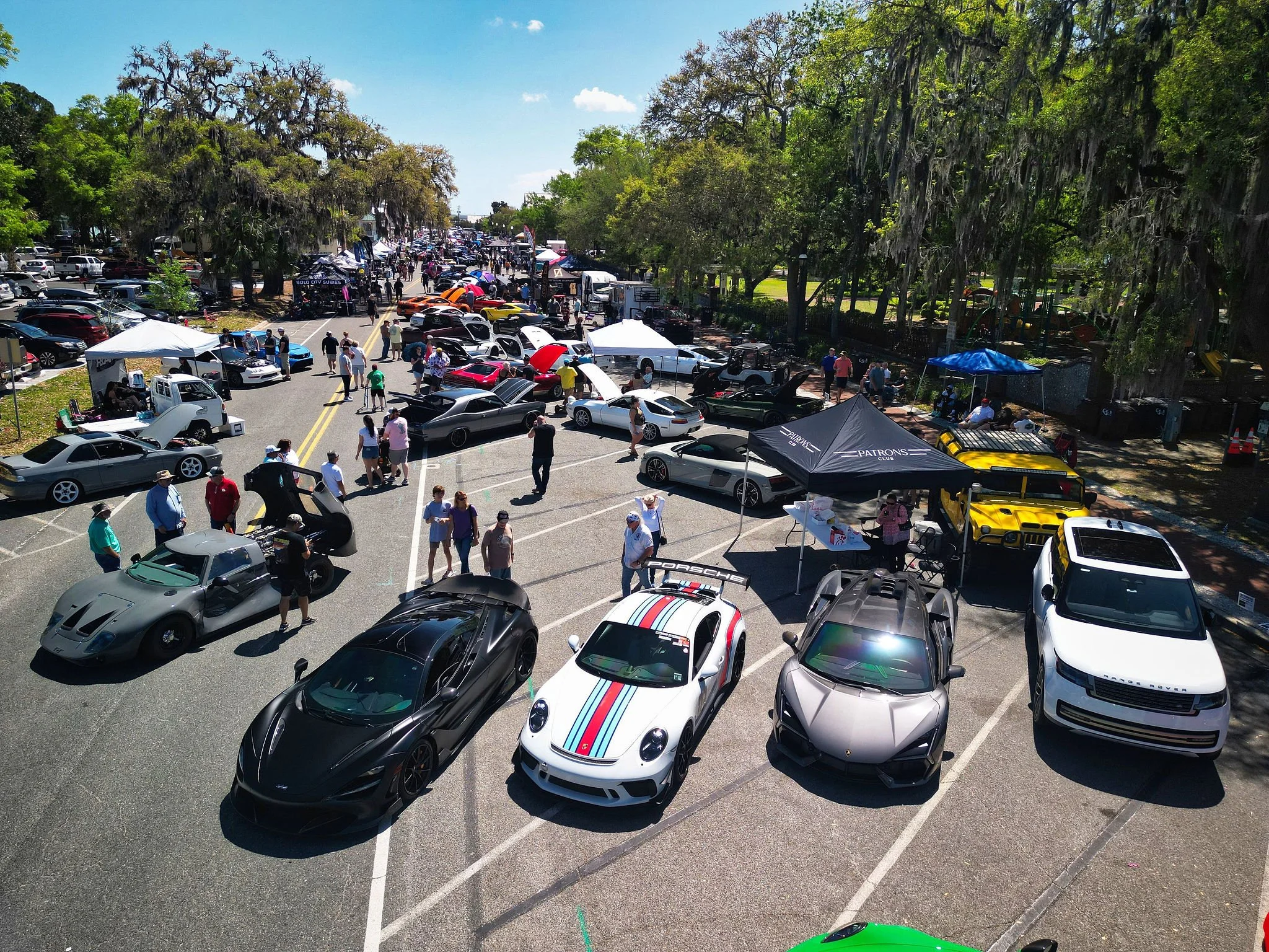 A car show in a parking lot with numerous luxury and sports cars, people walking around, and tents set up for vendors on a sunny day.