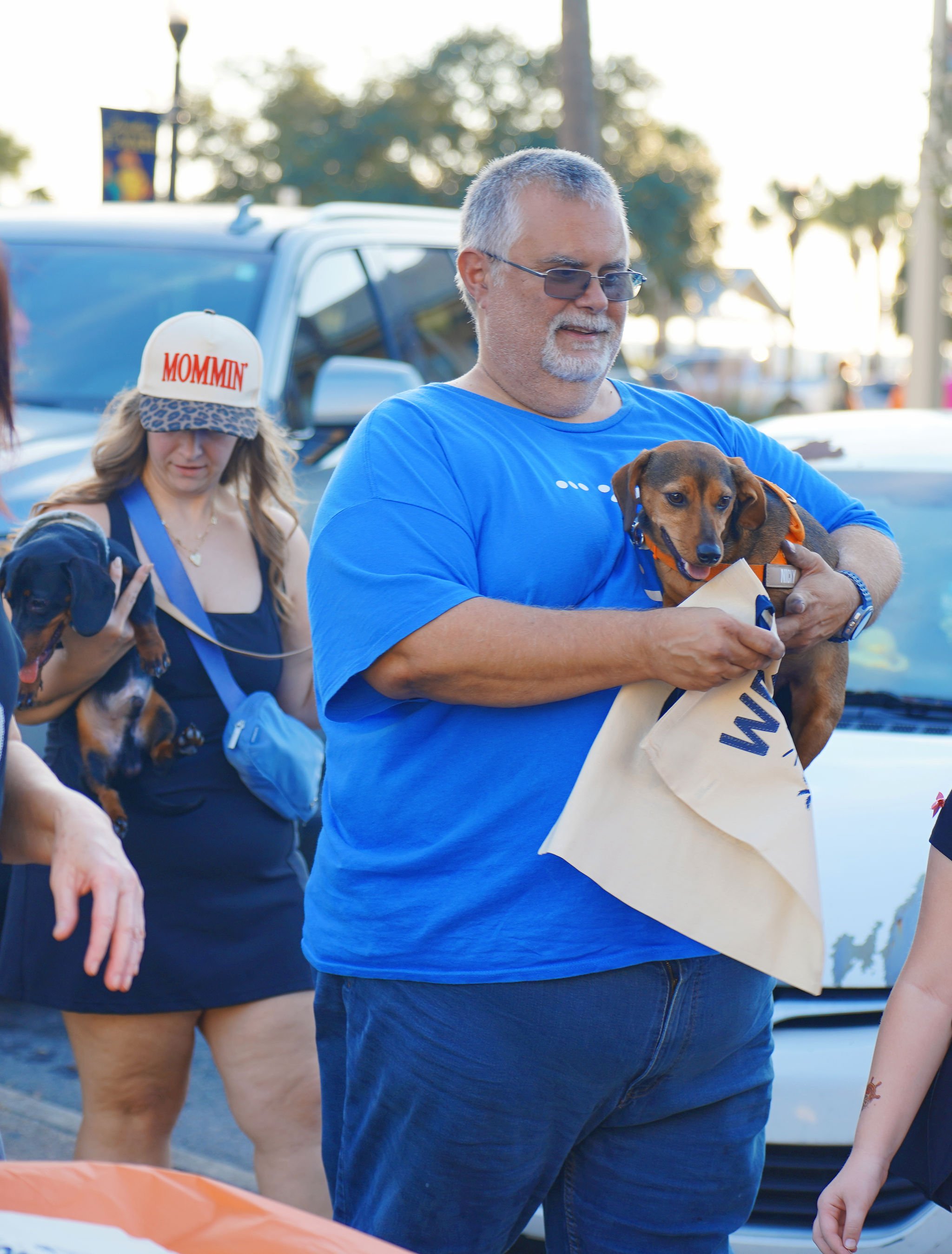 A man with gray hair, sunglasses, and a beard holds a small brown dog wearing an orange harness. The man is wearing a blue t-shirt and appears to be participating in a public outdoor event. In the background, a woman with long blonde hair, wearing a 