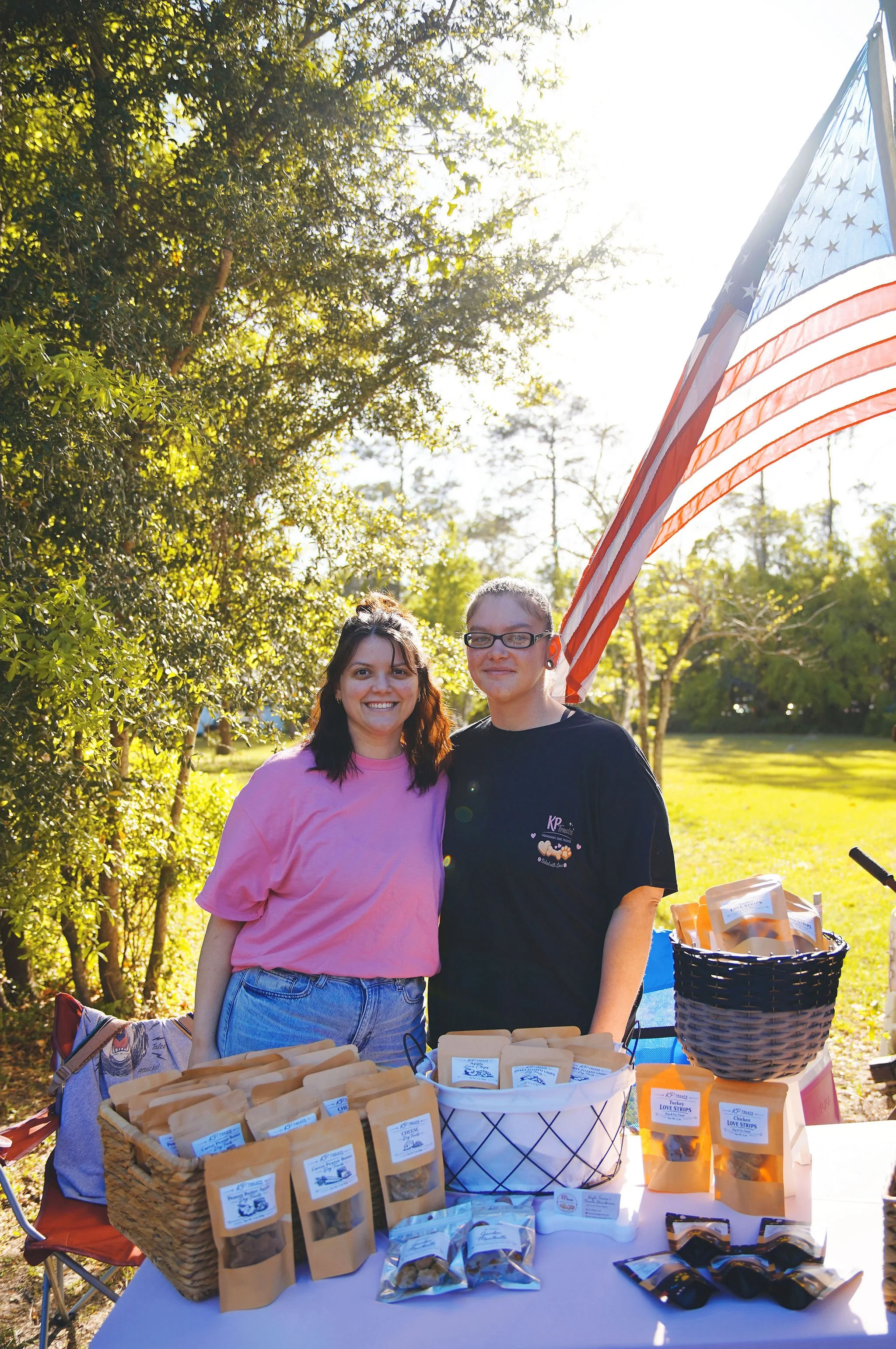 Two women standing outdoors behind a table with baked goods for sale, with trees and sunlight in the background, and an American flag.