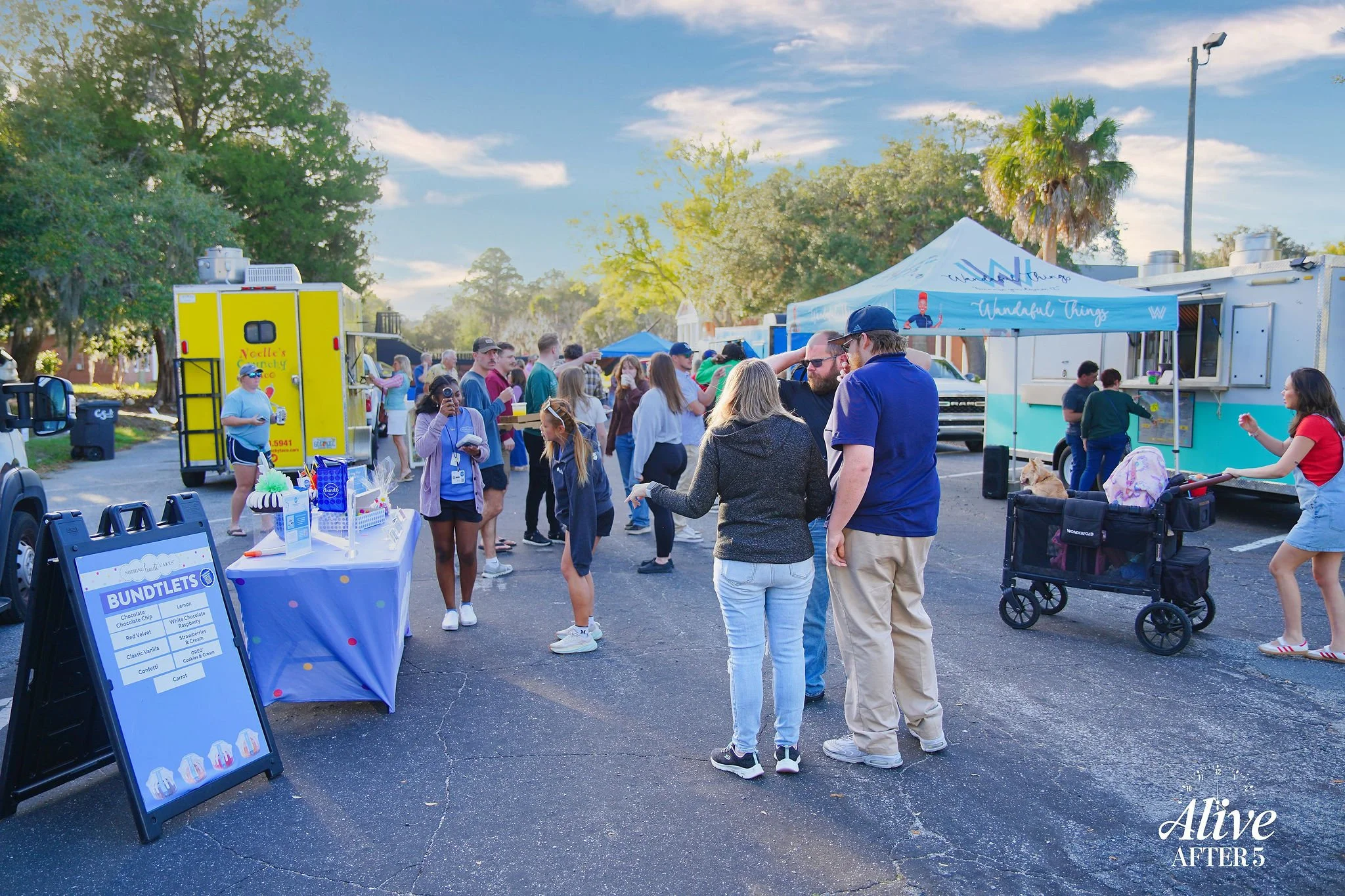People at an outdoor market or festival on a sunny day with vendors, food trucks, and tents, surrounded by trees and parked cars.