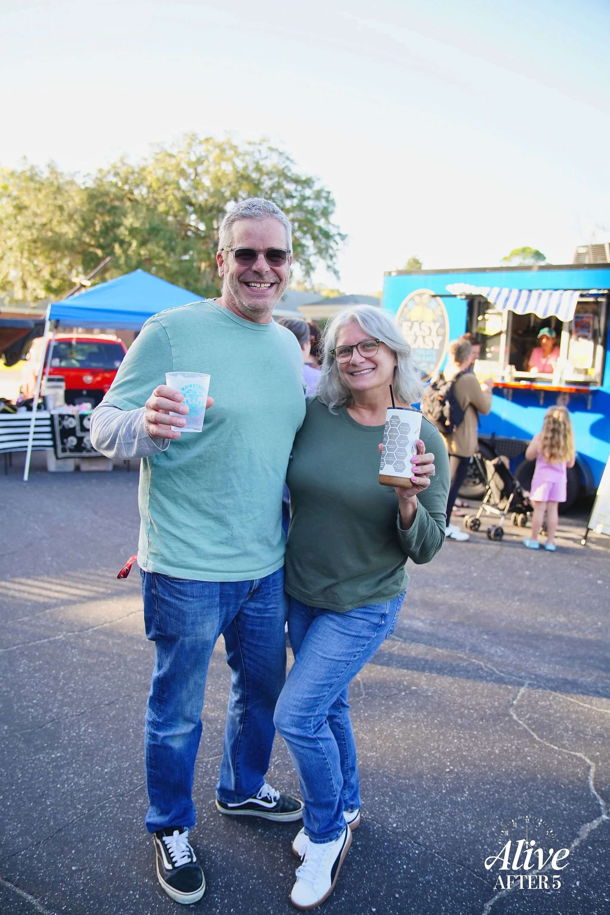 A smiling man and woman standing outdoors at a festival, holding drinks, with food trucks and market stalls in the background, wearing casual clothing and sunglasses.
