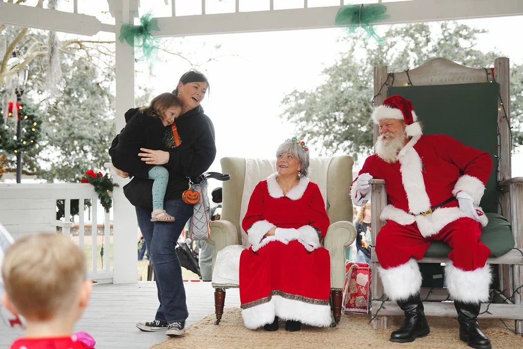 A woman holding a child in front of Santa Claus and Mrs. Claus during a Christmas event.