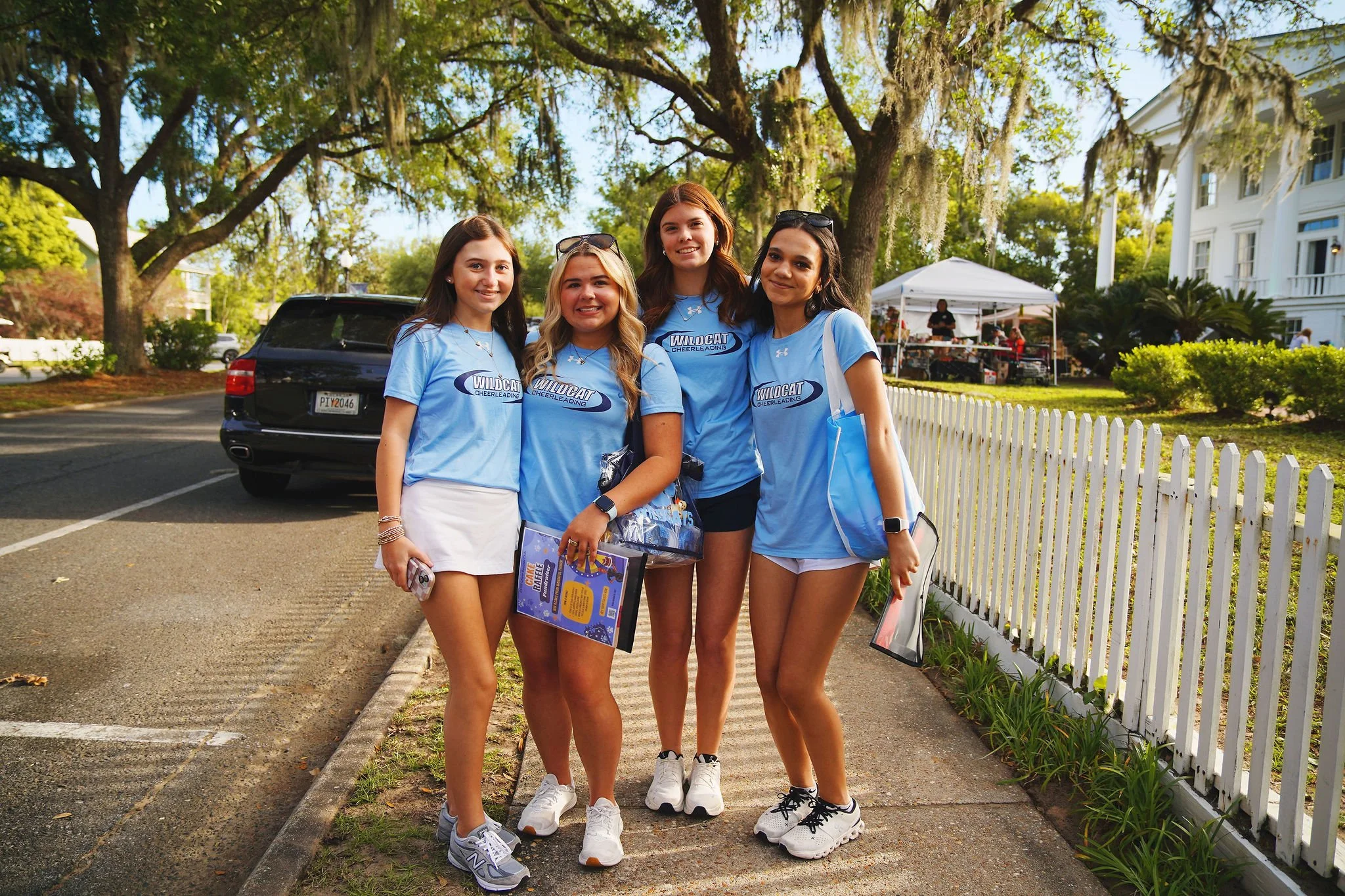 Four young women in light blue cheerleading shirts with ''Wildcat Cheerleading'' written on them, standing together on a sidewalk in a park-like setting, smiling at the camera.