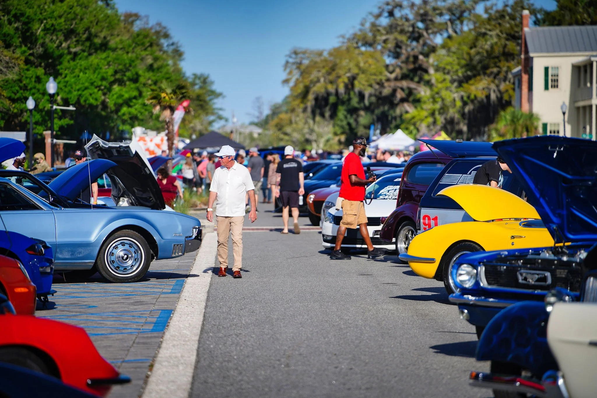 People attending a classic car show on a sunny day, viewing vintage cars displayed in a parking lot with trees and buildings in the background.