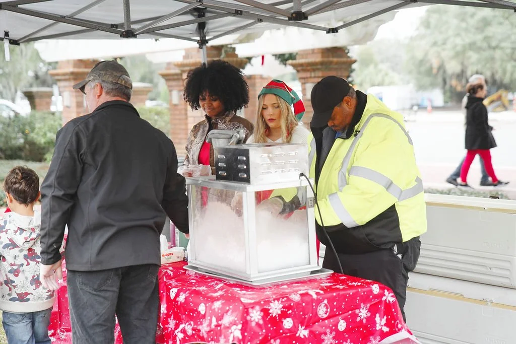People selling popcorn at an outdoor holiday event under a white canopy, with a red tablecloth with white snowflake patterns.
