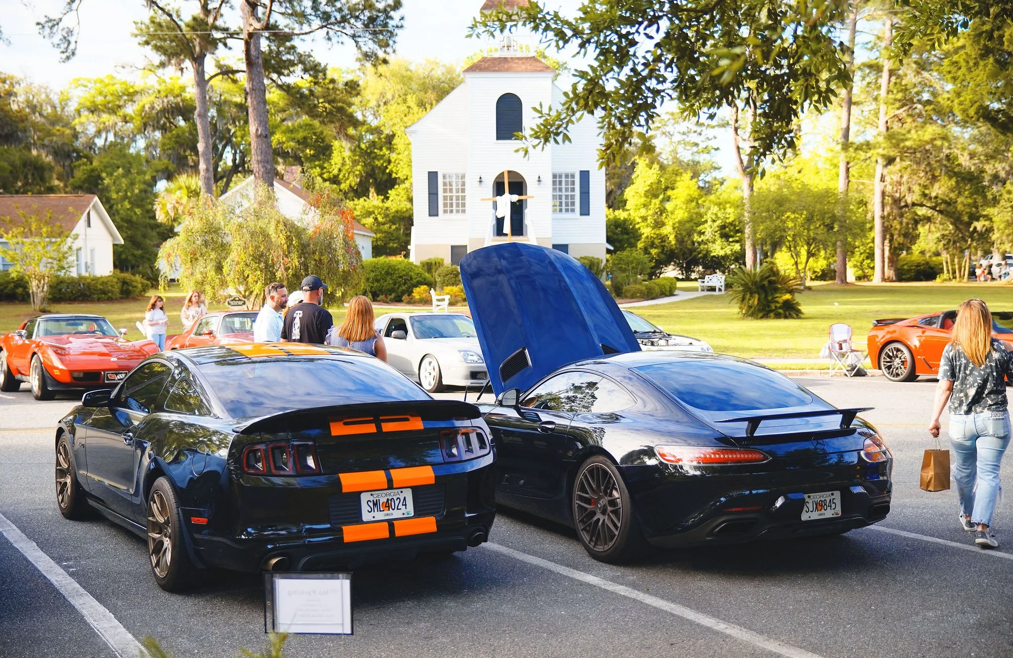 Multiple sports cars parked in a lot near a white church with people looking at the cars and talking, with trees and a sunny sky in the background.
