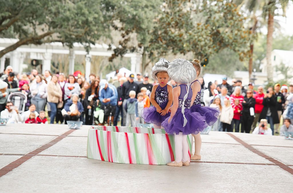Three young children dressed as elves with silver hats and purple tutus stand around a holiday-themed gift box on an outdoor stage, with a crowd of spectators in the background.