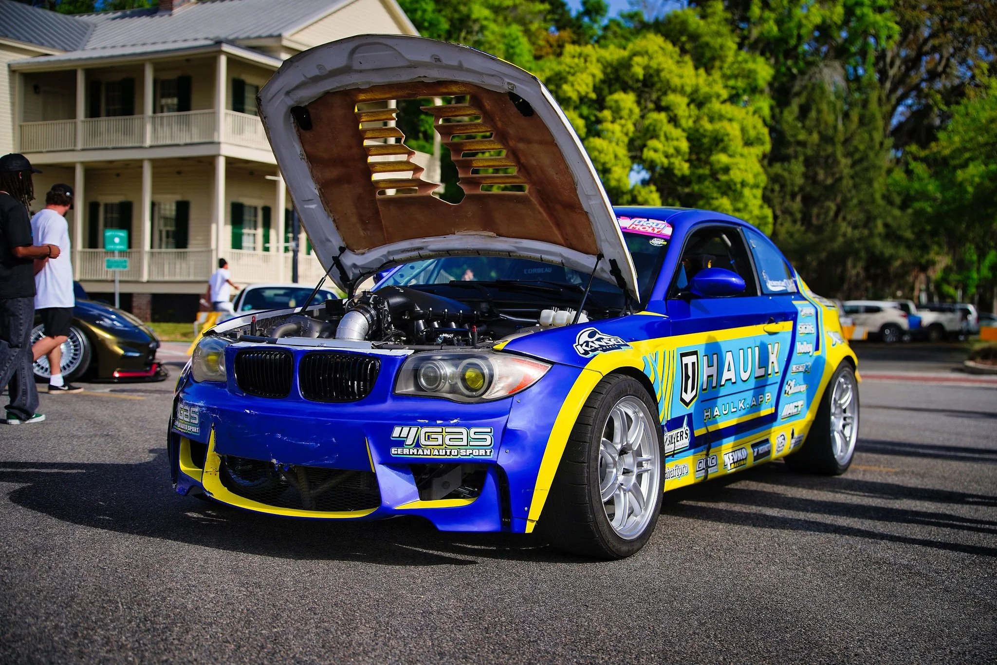 A blue racing car with yellow accents and sponsor logos, with its hood open, parked outdoors during a car event with people and other vehicles around.