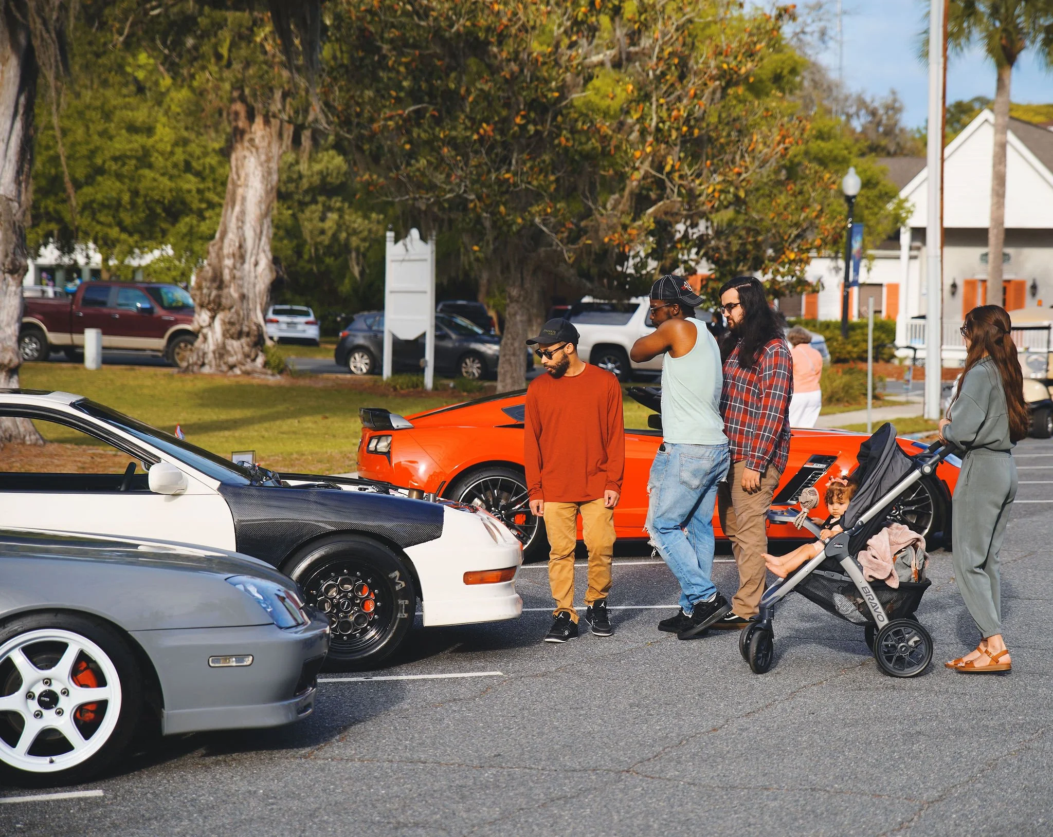 Group of people standing near parked cars in a parking lot with trees and houses in the background