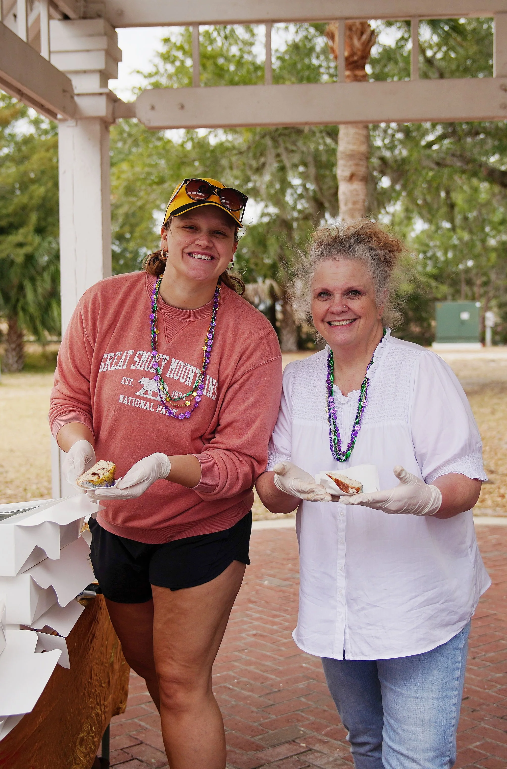 Two women wearing gloves and necklaces with beads, holding slices of pie and smiling at the camera outdoors under a white pavilion with trees in the background.