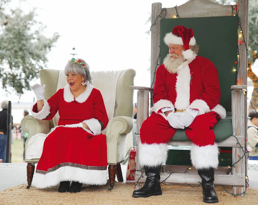 An elderly woman and man dressed as Mrs. Claus and Santa Claus sitting on chairs outdoors during a holiday event. The woman, wearing a red dress with white fur trim, gray hair and a holiday headband, is smiling and waving. Santa, with a white beard, 