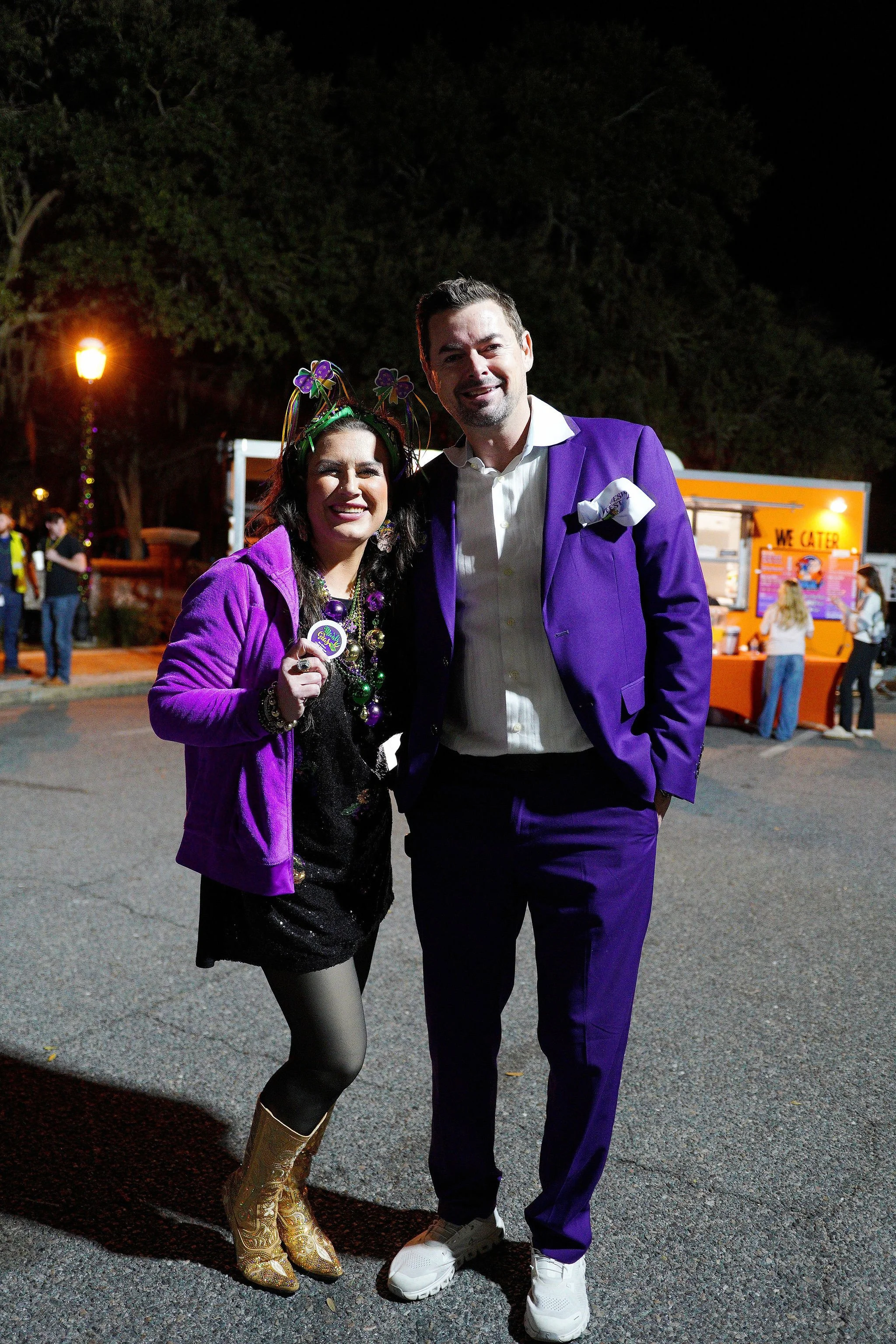 Two people dressed in purple at night, smiling and posing for a photo at an outdoor event, with a food truck and other attendees in the background.