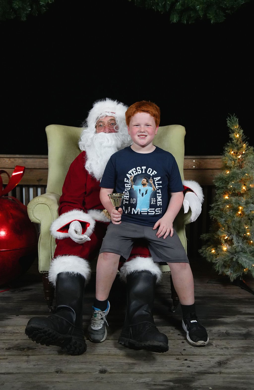 A young boy sitting on Santa Claus's lap, holding a small trophy, in a festive Christmas setting with a decorated tree on the right.