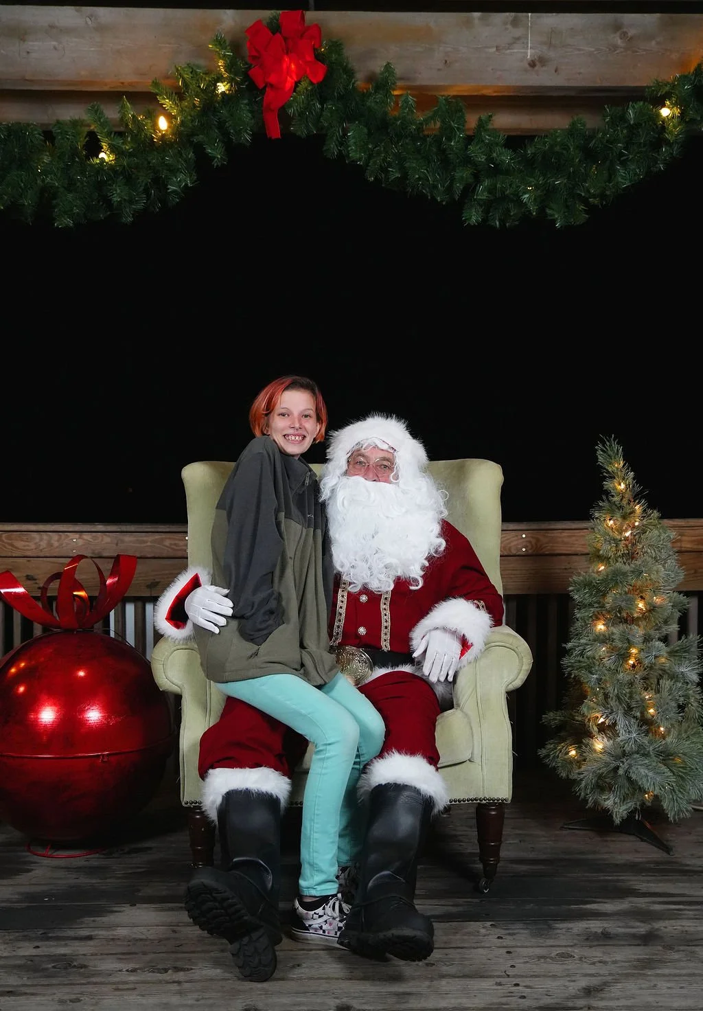 A girl sitting on Santa Claus's lap during a Christmas event. Santa is dressed in a traditional red suit with white trim, and the girl is smiling. The setting includes Christmas decorations like a large red ornament with a bow, a small Christmas tree