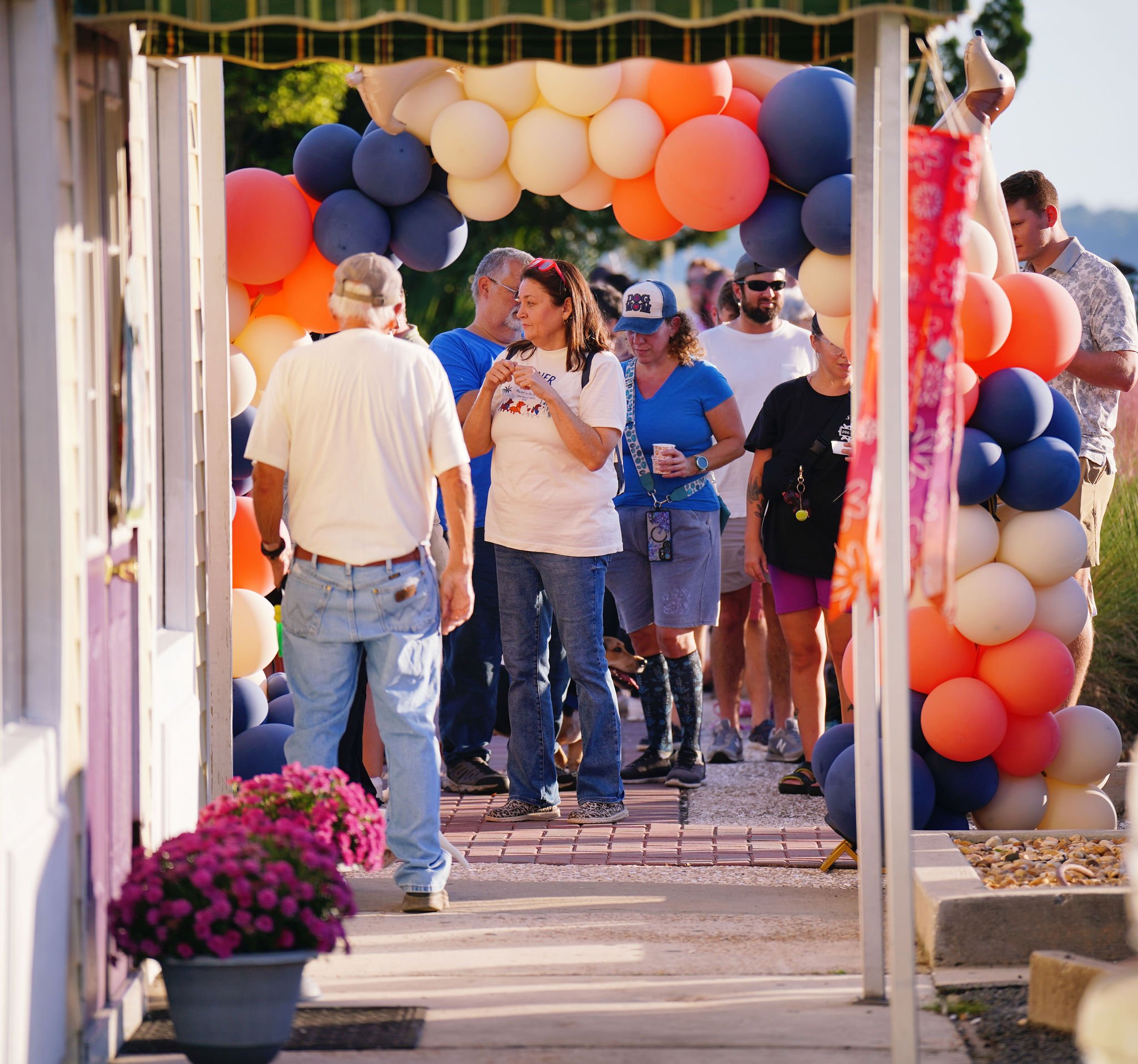 People gather under a decorated archway adorned with colorful balloons in shades of blue, cream, orange, and pink at an outdoor event, with trees and a clear sky in the background.