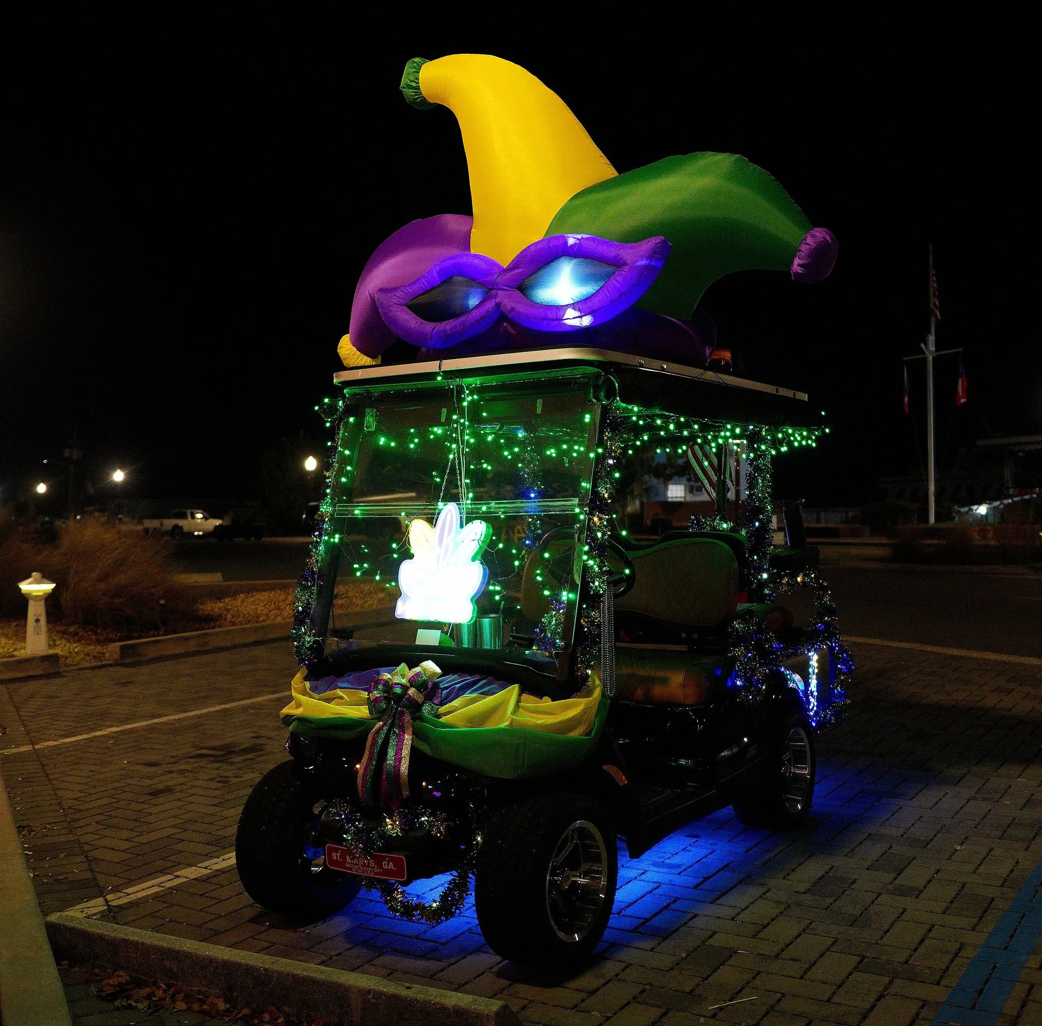 A decorated golf cart at night with Mardi Gras theme. It has a large inflatable hat and mask on top, and is adorned with colorful lights, tinsel, and a large ribbon bow.