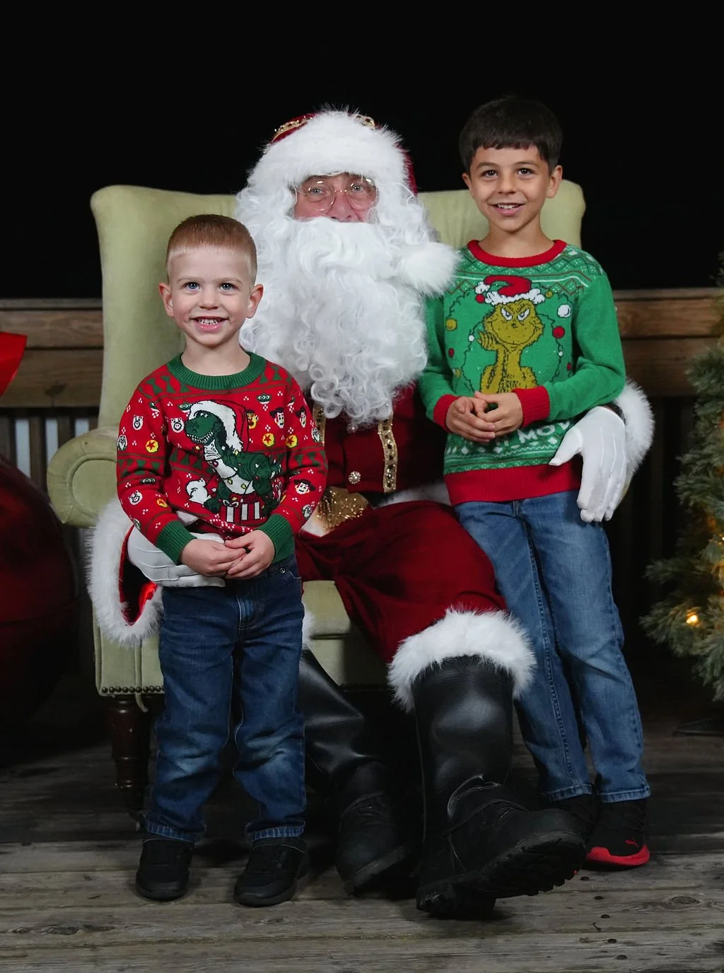 Two young boys sitting with Santa Claus, all smiling, wearing Christmas sweaters, in a festive holiday setting.