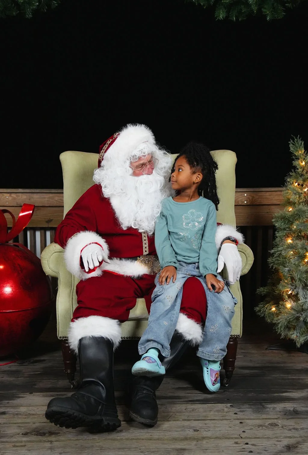 A young girl sitting on Santa Claus's lap, looking at him, in a Christmas setting with a decorated tree and gifts.