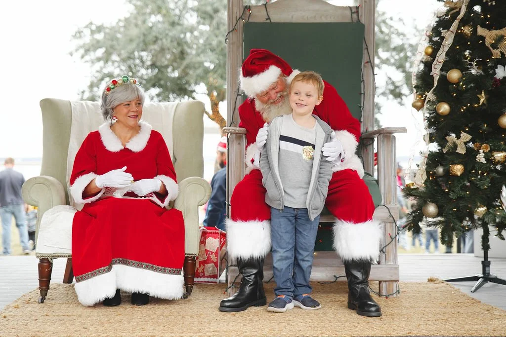 Santa Claus sitting on a throne with a young boy standing in front and a woman dressed as Mrs. Claus sitting nearby. There is a decorated Christmas tree beside them.
