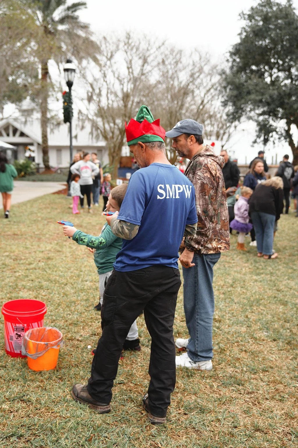 People gathered outdoors during daytime with trees and a white building in the background, some children and adults talking and playing, one man wearing an elf hat and a blue shirt with 'SMPW' on the back, and holding a toy gun.