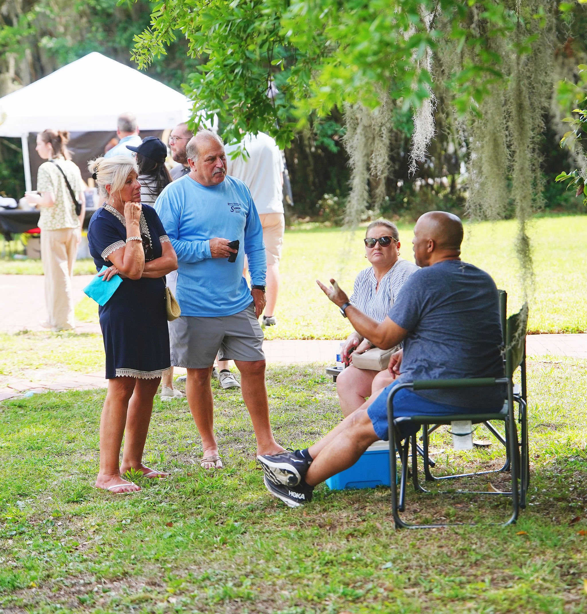 Group of people gathered outdoors, engaging in conversation under trees with Spanish moss, with a white event tent in the background, suggesting a social gathering or outdoor event.