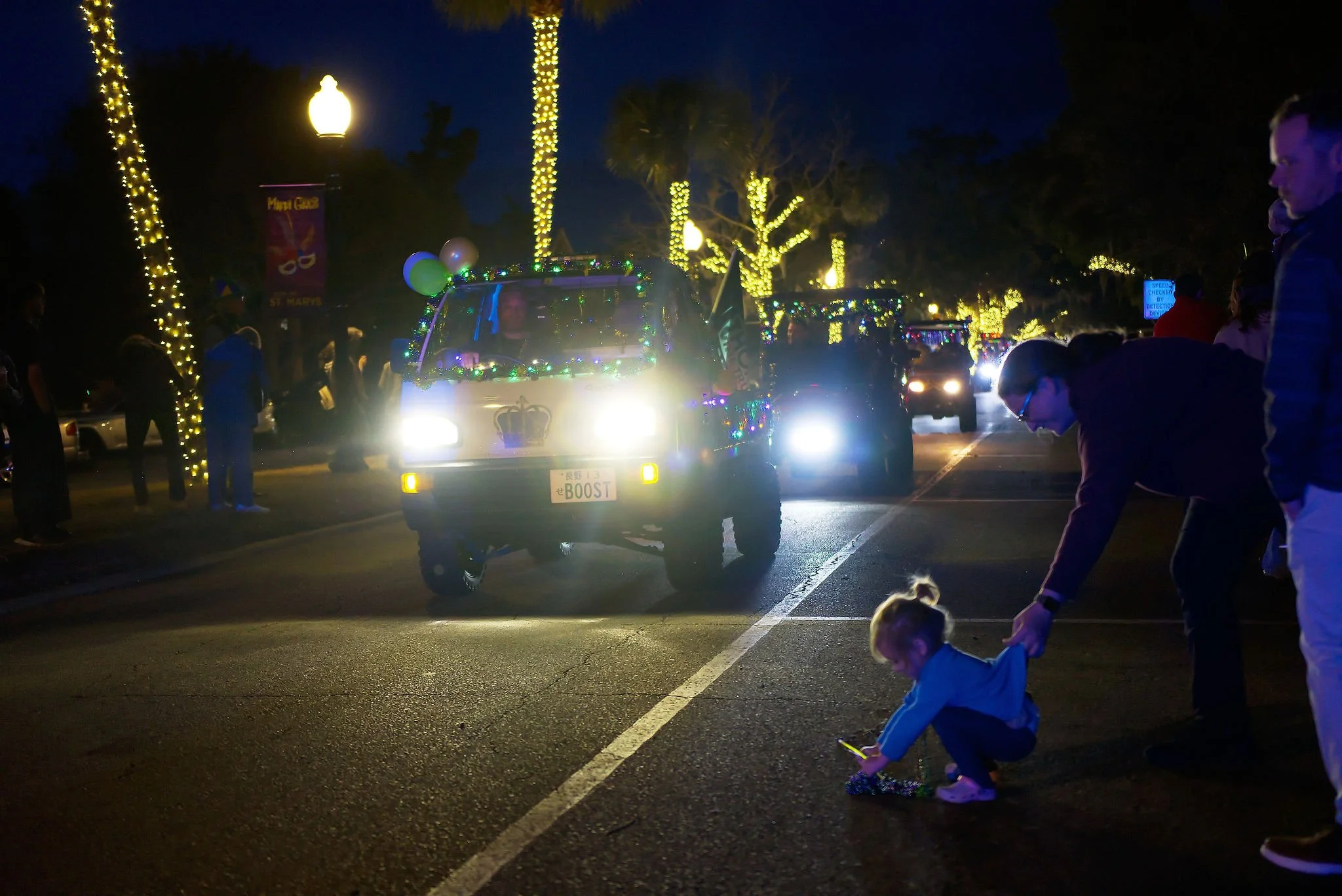 Nighttime parade with decorated cars, string lights on trees lining the street, people watching and children playing on the curb.