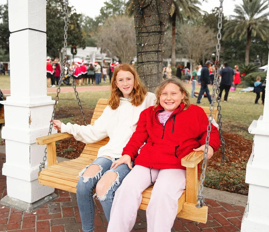 Two young girls sitting on a wooden swing with chains, smiling. One girl wears a white hoodie and distressed jeans, the other wears a red hoodie and light-colored pants. In the background, there are people in festive attire, trees, and holiday decora