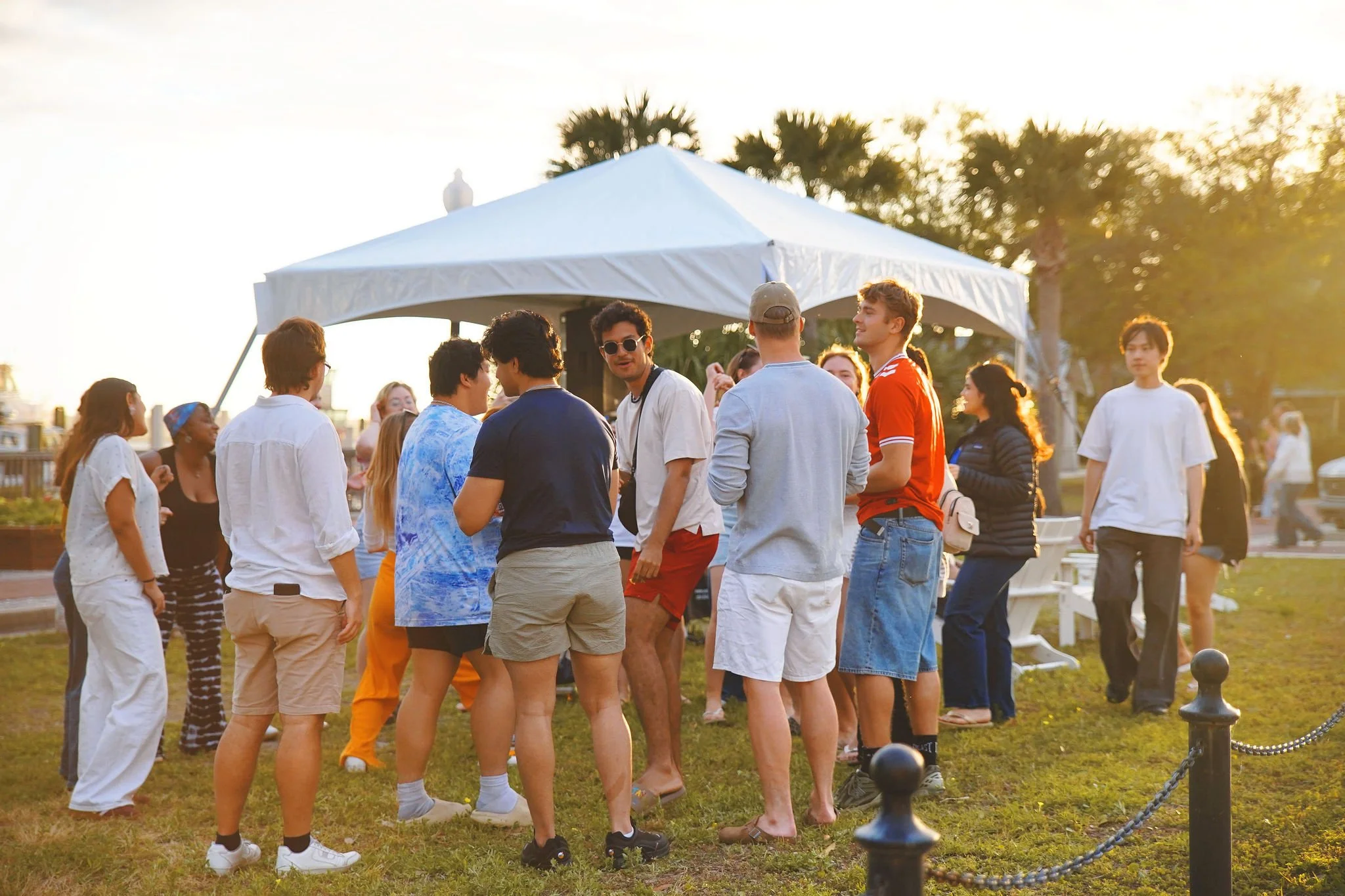 Group of people gathered outdoors during sunset, standing near a white canopy tent, engaging in conversations and enjoying a social event.
