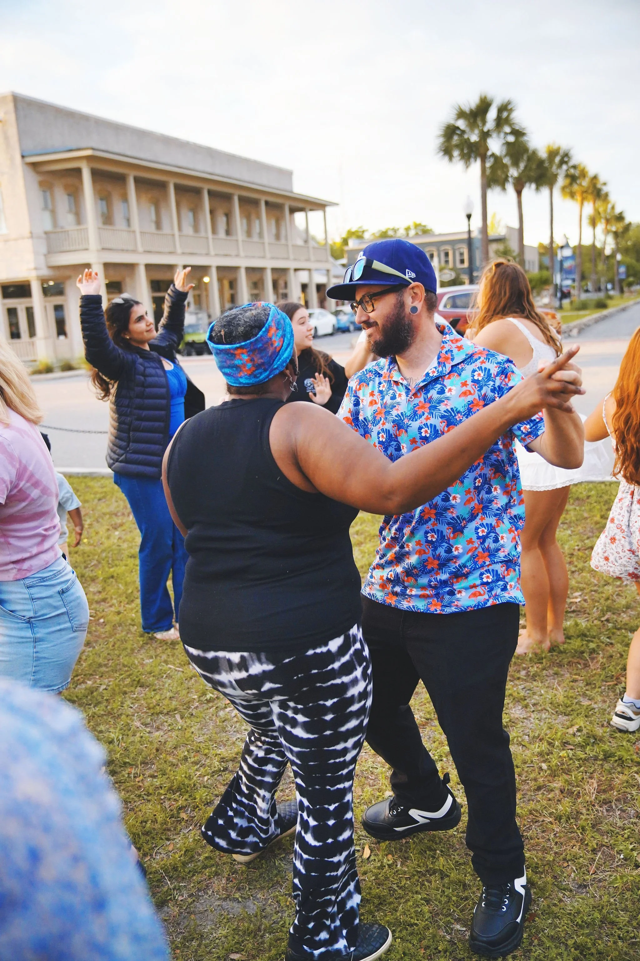 People dancing outdoors on a grassy area during a sunny day, with buildings and palm trees in the background.