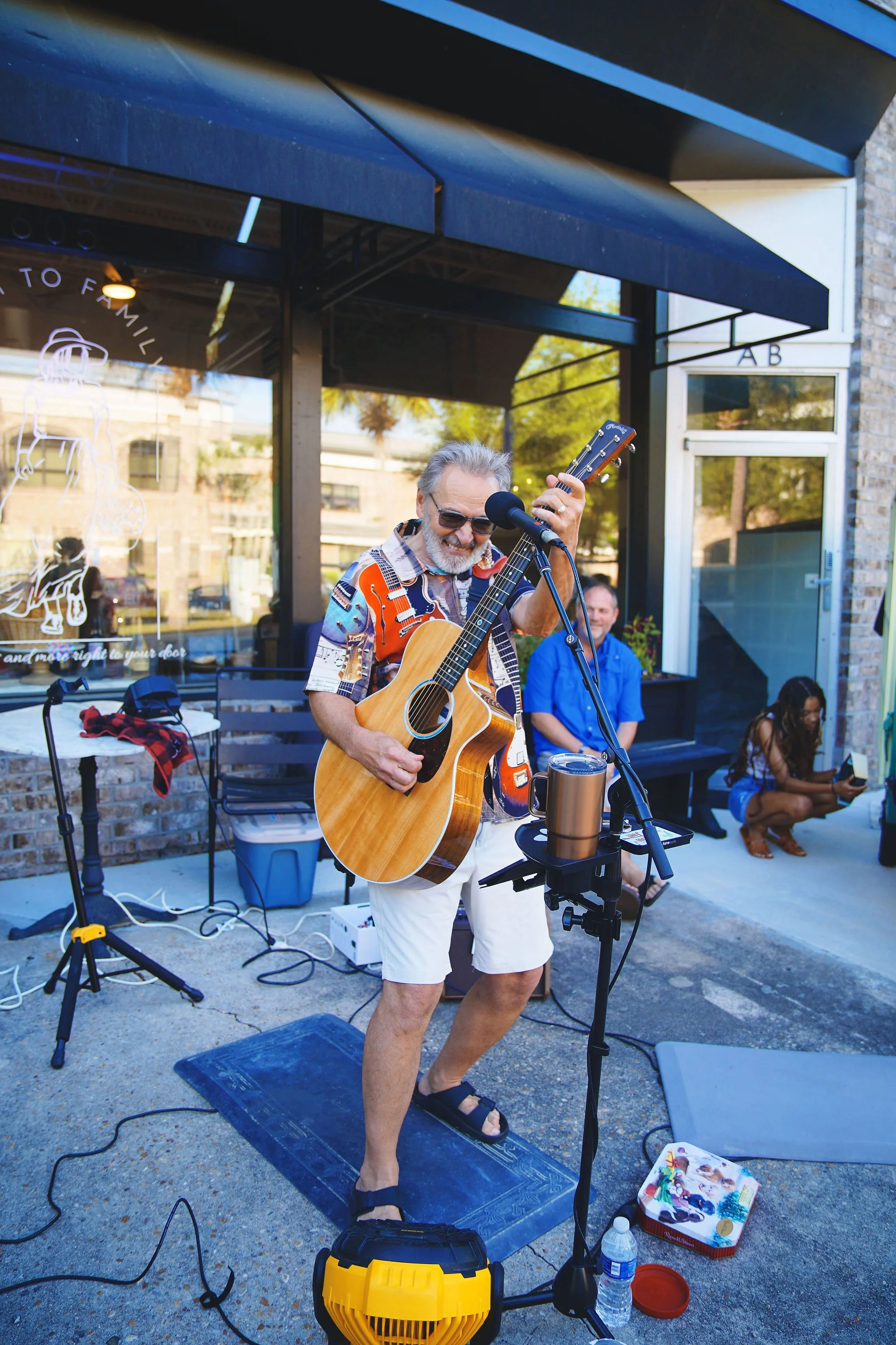 A man with gray hair and beard wearing sunglasses, a colorful short-sleeved shirt, and white shorts, playing an acoustic guitar and singing into a microphone outside a building. Two people are seated behind him, one in a blue shirt and the other in a