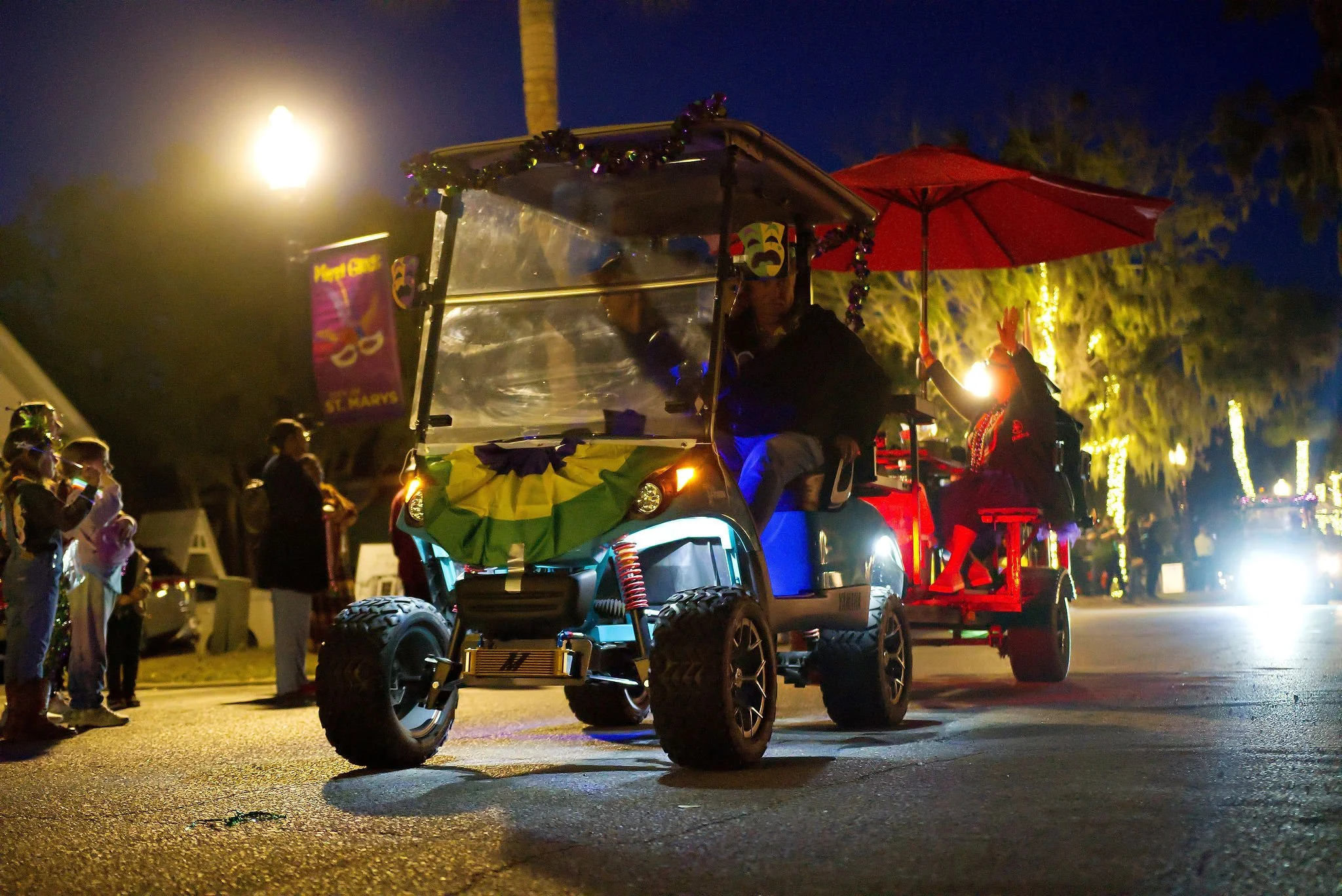 A decorated golf cart with people riding it, part of a parade at night, with onlookers on the sidewalk and festive lights in the background.