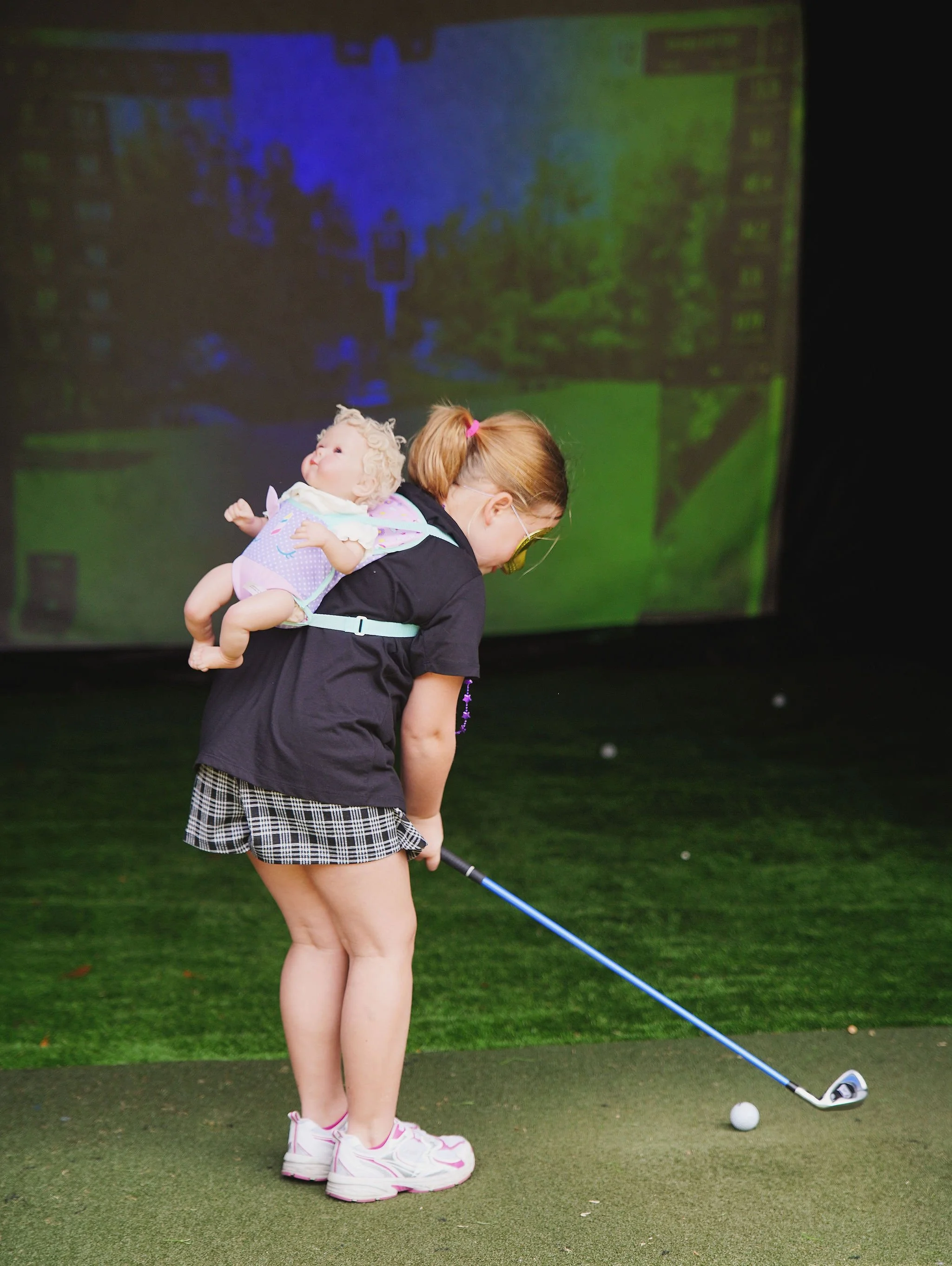 A girl with glasses and a ponytail practicing golf on a putting green, with a doll attached to her back. She is wearing a black shirt, checkered shorts, and pink sneakers.