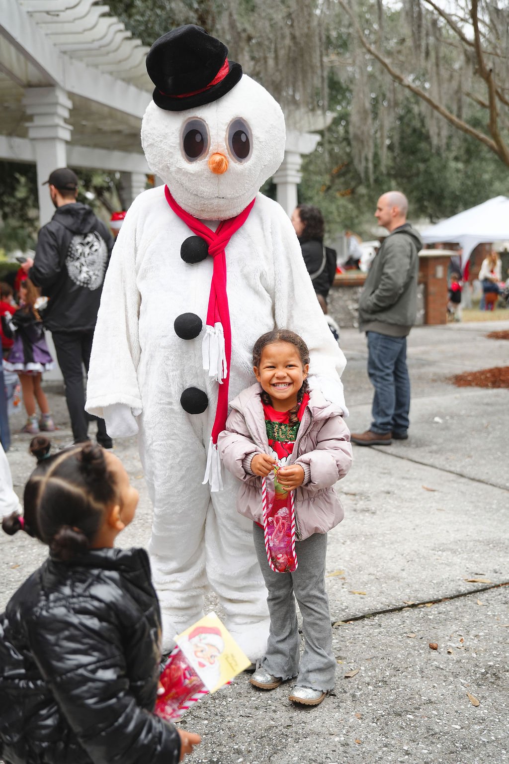 A young girl smiling happily wearing a pink jacket and holiday-themed shirt standing next to a snowman mascot with a black top hat, orange nose, and large eyes. The snowman is dressed with a red scarf and black buttons, and several people are in the 