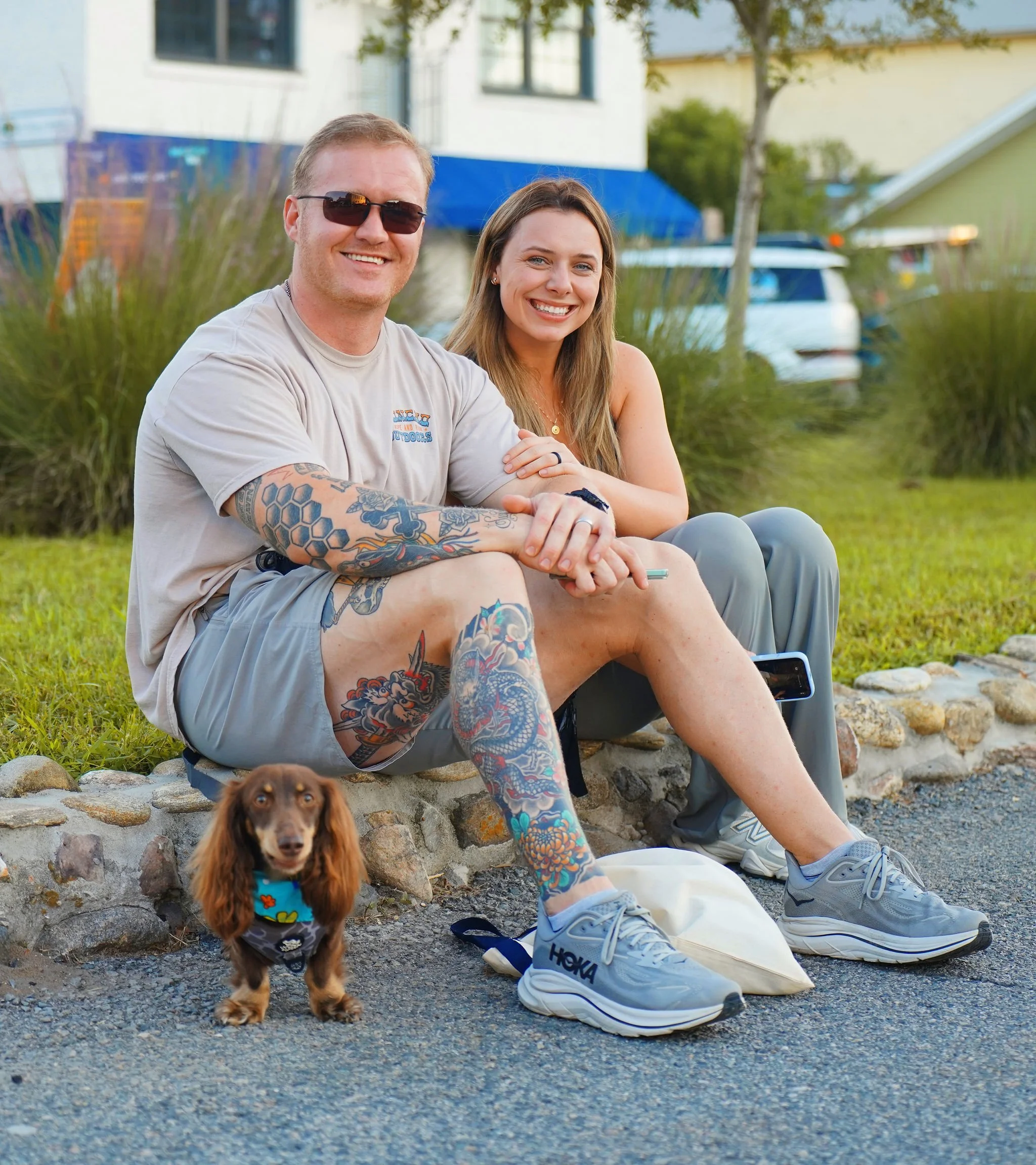 Two smiling people, a man wearing sunglasses and a woman, sitting on a stone wall with a small brown dog in front of them. The man has tattoos on his arms and the woman has long brown hair and is wearing a gold necklace. They are outdoors in front of